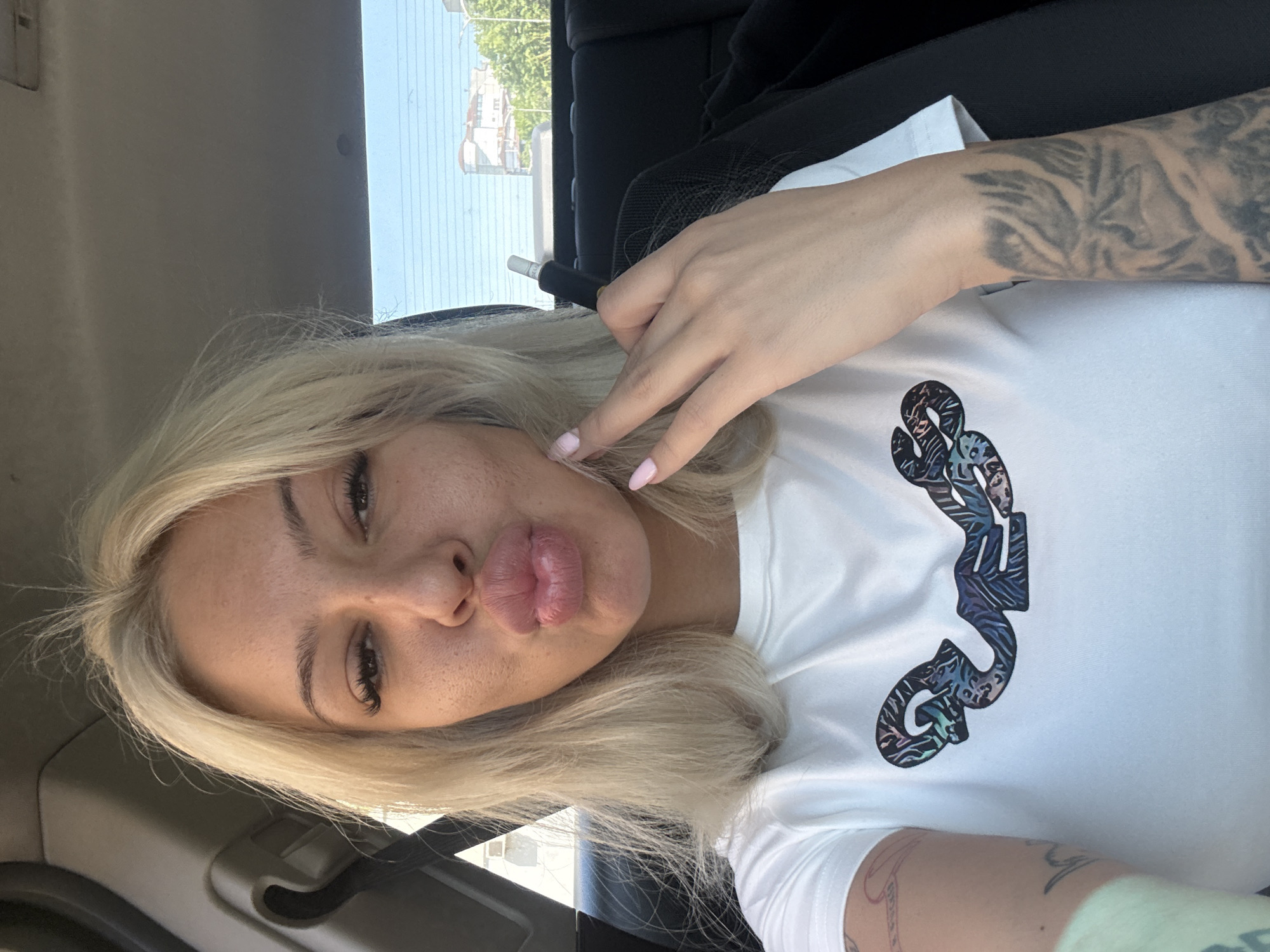A blonde-haired woman is featured in a close-up selfie taken inside a vehicle, likely a car. She is looking directly at the camera with a playful, pouty expression, her lips pursed. Her light skin shows some freckles, and her eyes are partially closed, emphasizing her long, dark eyelashes. Her light blonde hair is styled loosely around her face.
She wears a white short-sleeved t-shirt with a multi-colored, stylized graphic or logo on the chest, which is difficult to read but appears to have an abstract, almost camouflage-like pattern in blues, greens, and grays. Her right arm (on the left side of the image) is heavily tattooed with an intricate, dark pattern forming a full sleeve. Her left hand (on the right side of the image) is raised near her face, with two fingers extended, and her nails are painted a light pink. She holds a small, black and white cylindrical object, possibly a vape pen, between her fingers.
Through the window in the background, a sunny outdoor scene is visible, showing clear skies, green trees, and glimpses of distant buildings, suggesting an urban or suburban setting. The bright light streaming in indicates it is daytime. The interior of the car is visible, including a dark seatbelt and a light-colored headliner. The location context provided is Plovdiv, Bulgaria.
