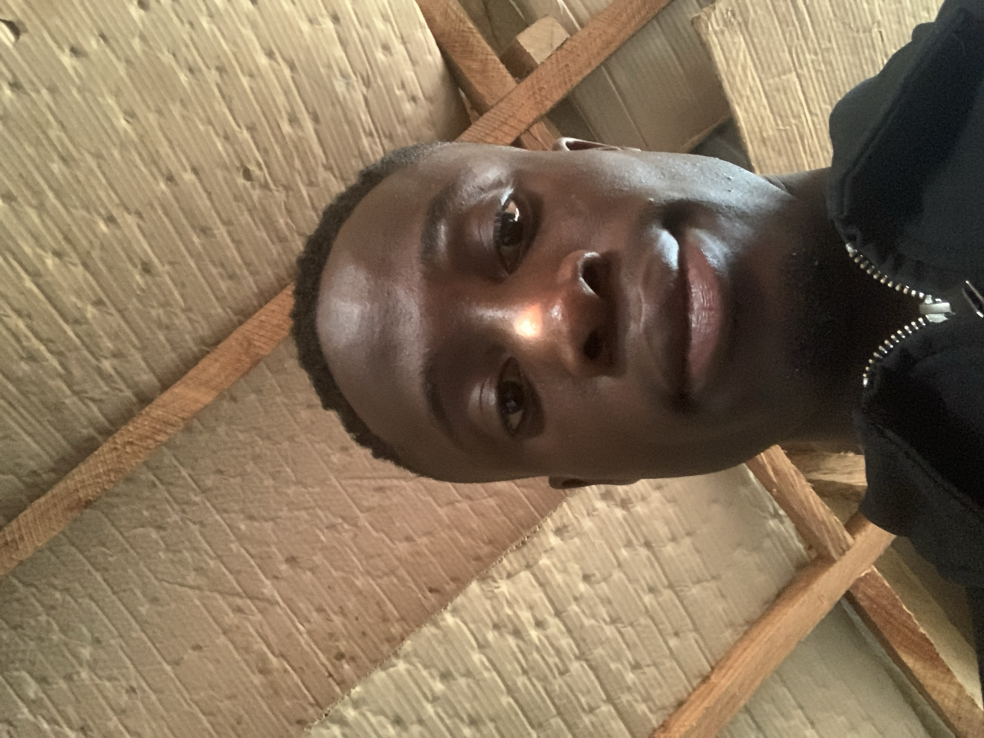 A close-up, low-angle selfie of a dark-skinned man wearing a dark jacket with a visible silver zipper. The man is smiling. The background is a ceiling constructed with wooden beams and textured, light-colored material, potentially insulation or cardboard. The photograph was likely taken indoors. The location in Gbeleko, Nigeria, is unknown based on the visual data. No specific actions or interactions are discernable.