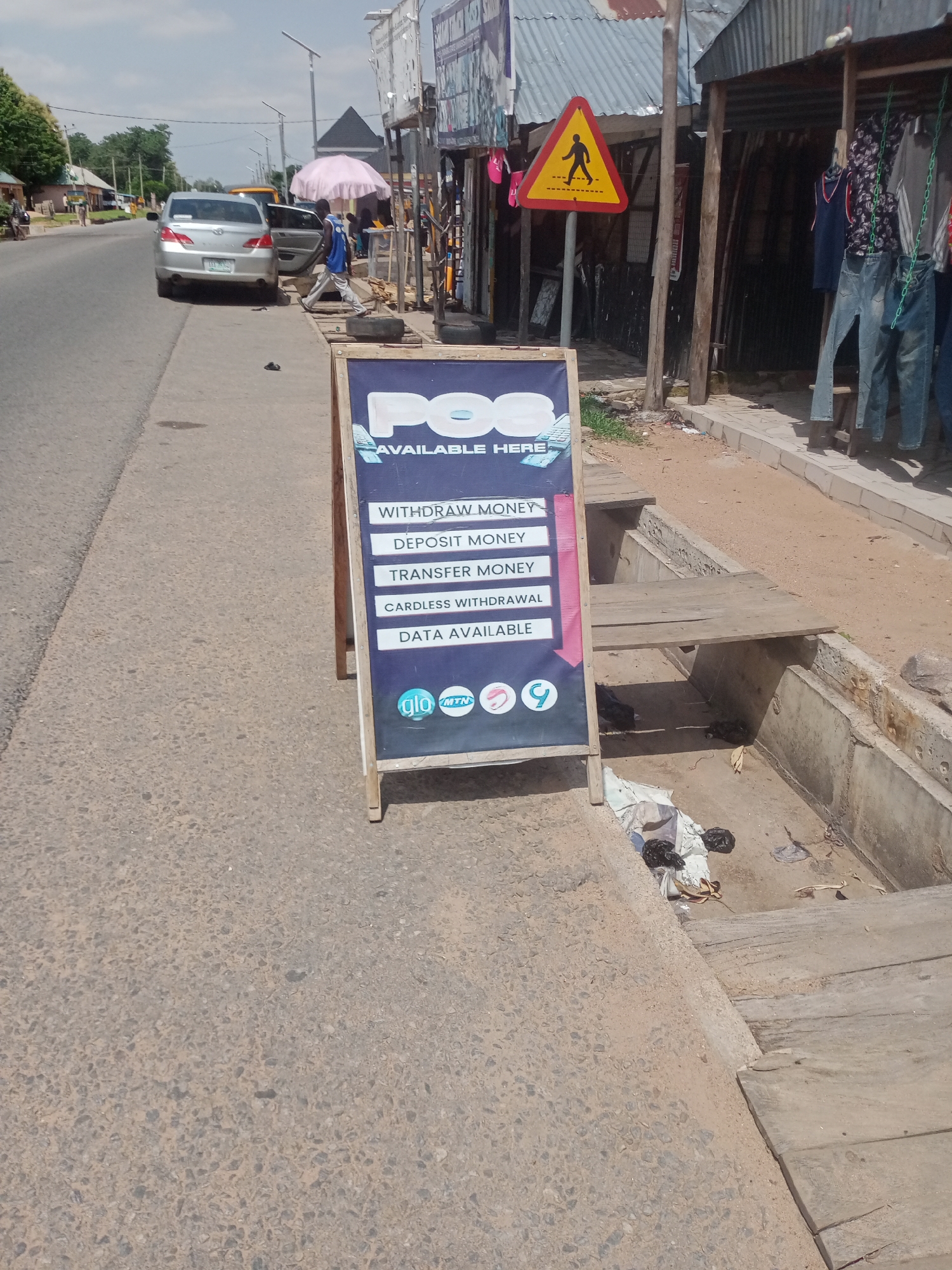 This media file captures a street scene in Jalingo, Nigeria, on a bright, sunny day. The foreground is dominated by a dark blue A-frame sign advertising "POS AVAILABLE HERE." The sign lists various services offered, including "WITHDRAW MONEY," "DEPOSIT MONEY," "TRANSFER MONEY," "CARDLESS WITHDRAWAL," and "DATA AVAILABLE." Below these options, logos for mobile network providers "glo" and "MTN," along with another circular logo, are visible.
To the right of the sign, a concrete drainage channel runs alongside the road, partially covered with wooden planks. Some scattered debris and refuse are visible within the channel. In the background, a silver car is parked on the side of a paved road. A person in a blue shirt and grey trousers is seen standing next to the open driver's side door of the car. Further down the road, other individuals and structures, including a pink umbrella and a yellow pedestrian crossing sign, suggest a bustling commercial or residential area. Clothing is displayed for sale outside a shop on the right side of the frame. The overall impression is of a typical day in a Nigerian town, with everyday commerce and infrastructure in view.