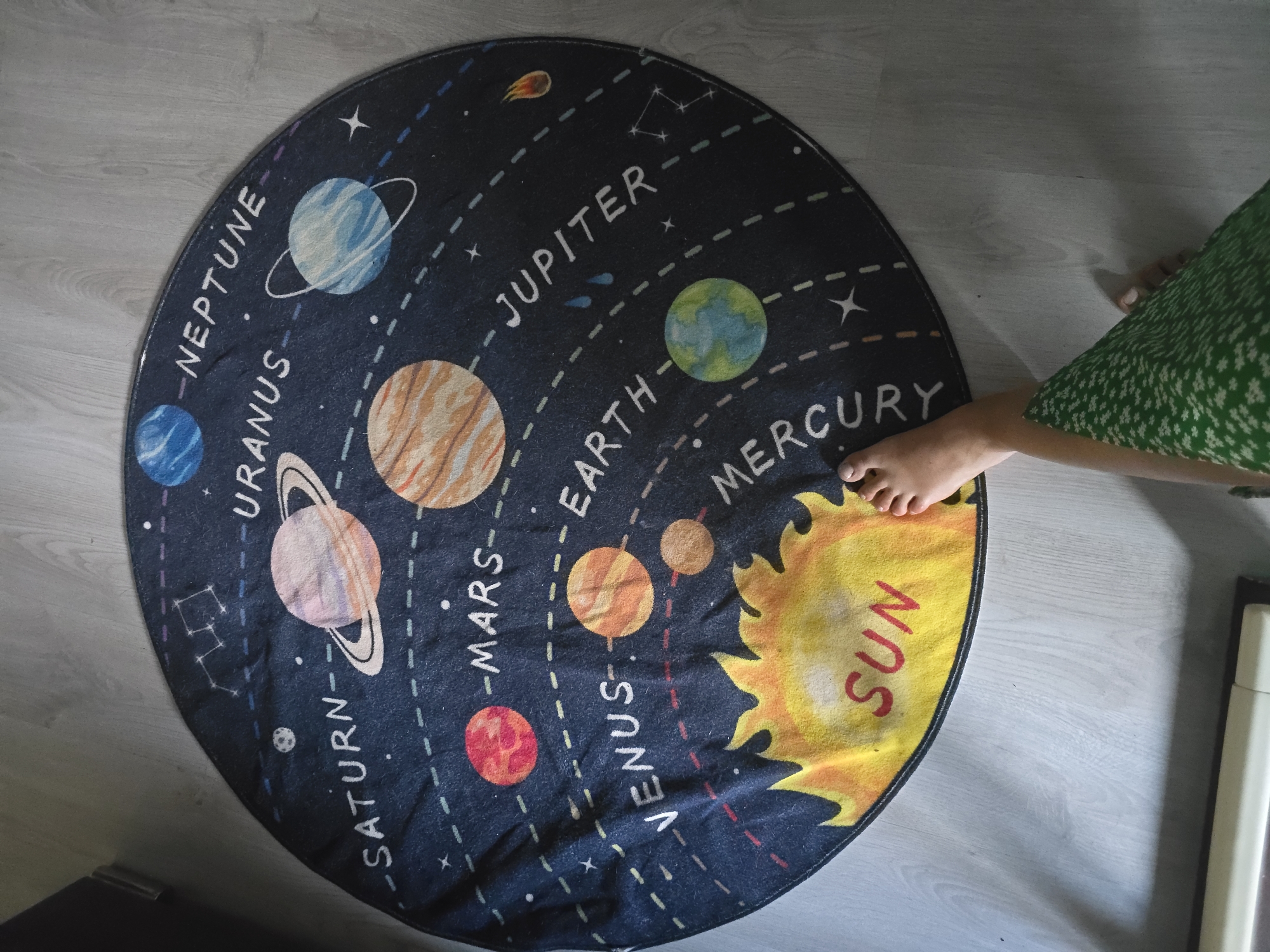A round rug depicting the solar system is spread on a light wooden floor in Golyanovtsi, Bulgaria. The rug features illustrations of the sun and planets, including Mercury, Venus, Earth, Mars, Jupiter, Saturn, Uranus, and Neptune, along with stars and constellations. A person's bare feet, one with painted toenails, are visible on the right side of the rug, with a green patterned fabric draped over their leg. The lighting suggests it is daytime, with shadows cast on the floor.