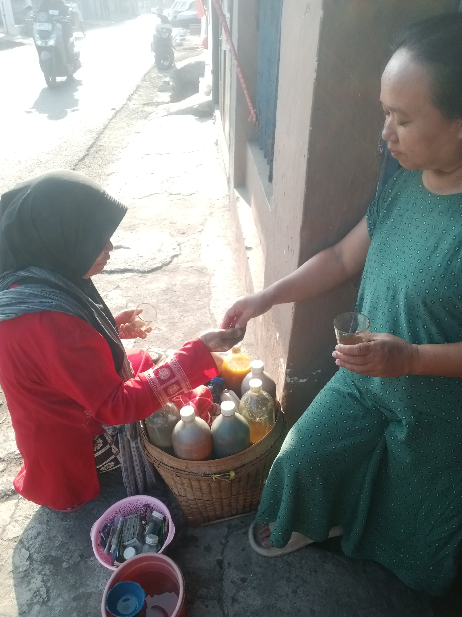 Two women are interacting on a street in Tasikmalaya, Indonesia. The woman on the left wears a red top and a black headscarf. She is holding a small clear cup and a small object, possibly a coin, in her hands. She is facing a basket filled with various bottles and containers, some appearing to contain liquid. She is leaning towards the basket as if selecting an item or making a transaction. The woman on the right wears a green patterned dress and is holding a glass of amber-colored liquid. She is looking down and her arm is extended towards the basket. The setting appears to be a street scene with a building wall to the right. There are motorcycles and a car visible in the background, suggesting a public thoroughfare. The ground is paved and there are shadows indicating sunlight. In the foreground, there are two buckets, one pink and one red, with a blue cup in the red bucket.