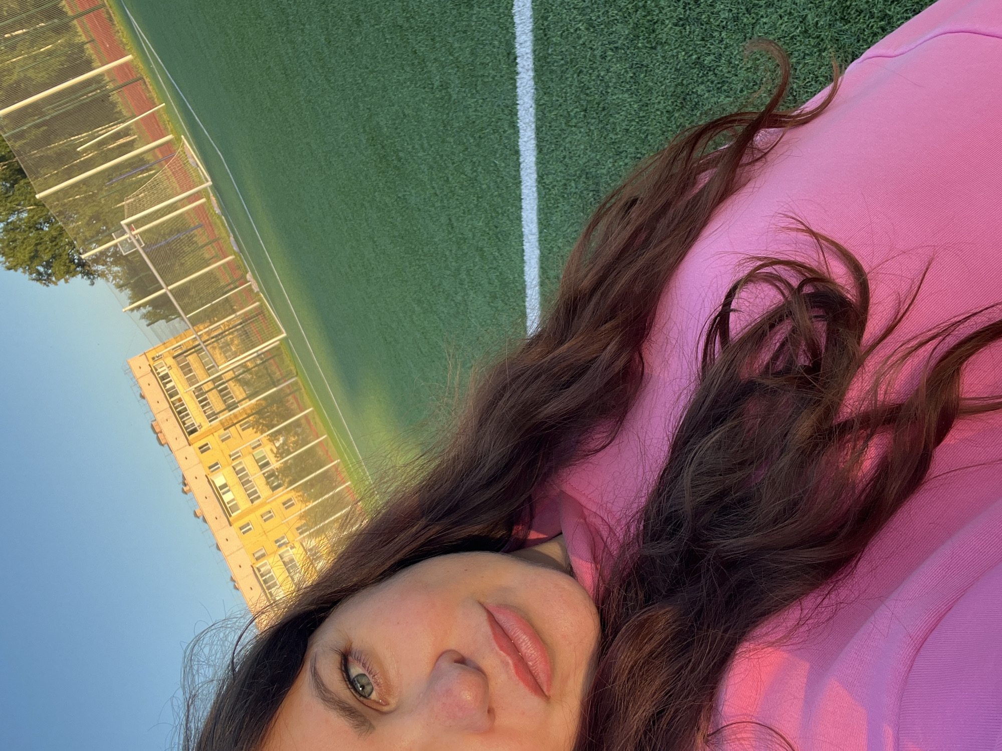 The image is a close-up selfie taken outdoors, with the subject, a woman, lying on her side on what appears to be a green artificial turf field, possibly a sports field or playground. She is wearing a pink hooded sweatshirt. Her long, dark brown hair is spread across her shoulders and the sweatshirt. The woman's face is partially visible, with her eyes open and looking towards the camera, and her lips slightly parted and colored with lipstick.
In the background, the field has a white line marking its edge. Beyond the field, a tall, light-colored building with many windows is visible. There are also trees and a metal fence structure that appears to be part of a sports facility, possibly a soccer goal. The sky is a clear, bright blue, suggesting it is daytime and the weather is good. The lighting indicates it might be late afternoon or early evening due to the warm hue on the pink sweatshirt.
The overall impression is casual and relaxed, possibly taken during a moment of leisure or sport. There is no visible text in the image.