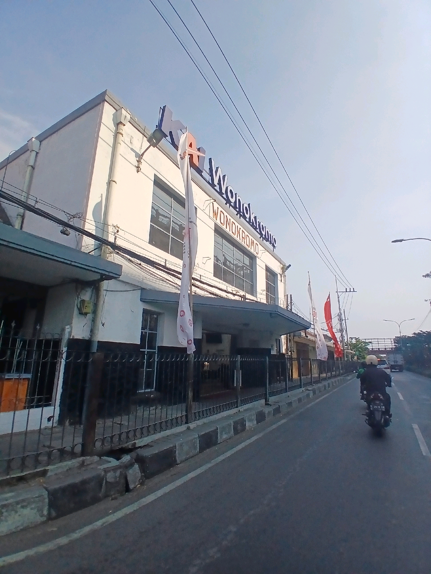 The image shows a street scene in Surabaya, Indonesia, on a clear day with a bright sky. On the left side of the road is a white building with large windows and prominent blue signage reading "KA WONOKROMO". Two flags are displayed on poles in front of the building; one is white with red and white circular emblems, and the other is red. A black metal fence runs along the front of the building.
The road in the foreground is paved and has a white line marking the lane. A person is riding a motorcycle away from the camera in the right lane, wearing a black jacket and a white helmet. The motorcycle has a license plate that reads "S 2513 LX". In the background, other vehicles, including a truck, can be seen further down the road. The environment appears urban, with trees and streetlights visible along the roadside.