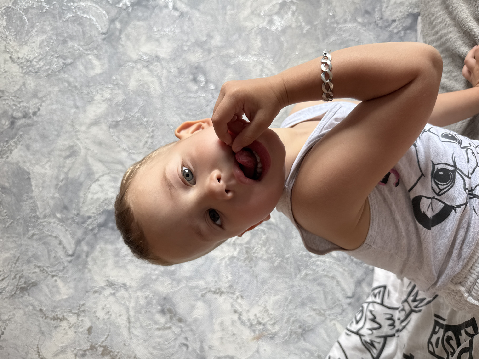In a medium shot, a young child with light brown hair and blue eyes is seated indoors, looking directly at the camera with their mouth open and tongue extended. The child is wearing a grey tank top with a graphic print and a silver bracelet on their left wrist. Their left hand is touching their cheek near their mouth. The background consists of a textured grey wall and a portion of a bed with a black and white patterned sheet. The scene is likely within a residential setting in Rusanovo, Russia.