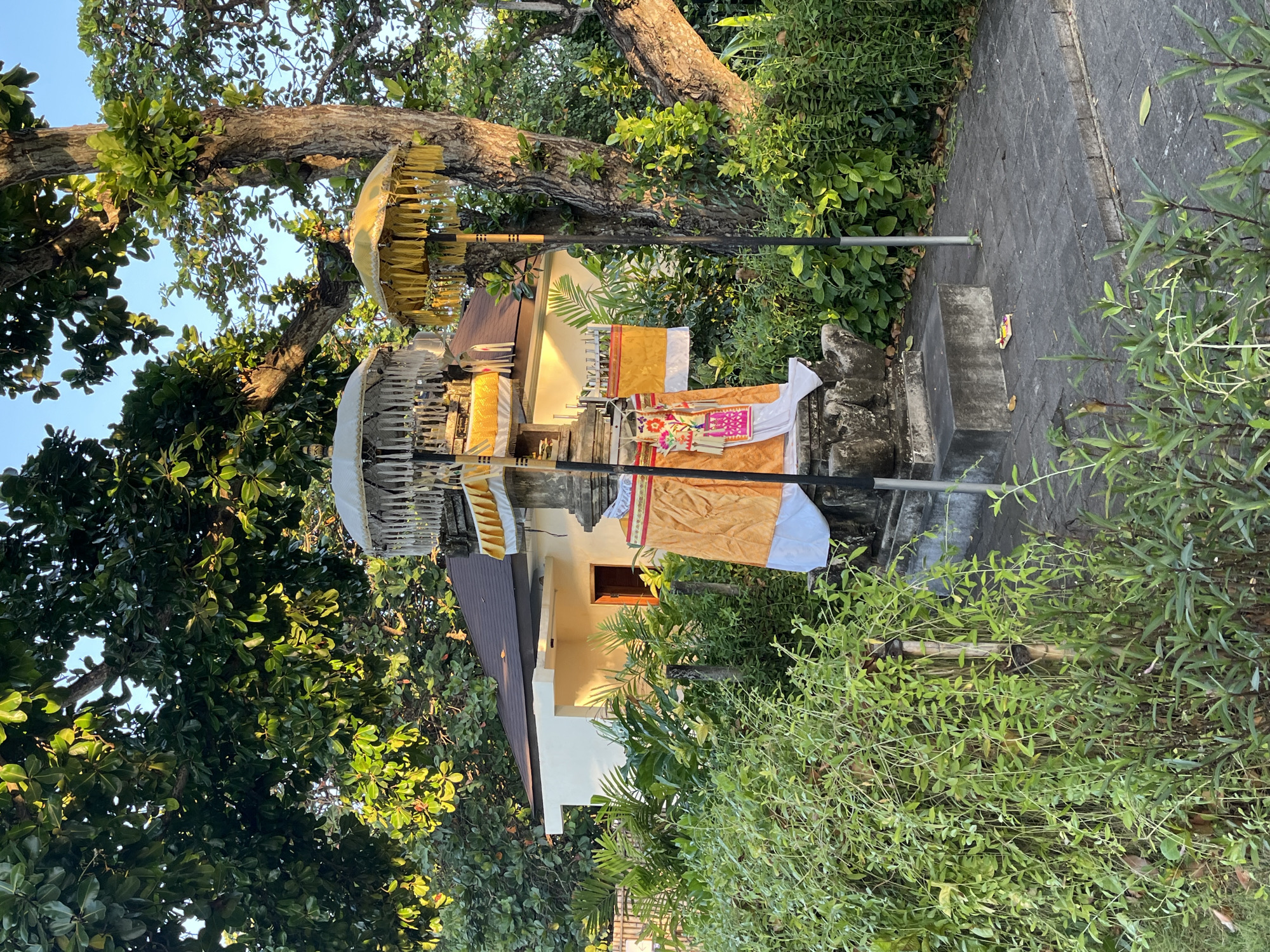 A stone structure, likely a shrine, is adorned with ornate umbrellas and decorative fabrics, including gold and white cloth, and a colorful embroidered banner. The shrine sits on a paved walkway, surrounded by lush green foliage and trees. In the background, a modern building with a brown roof is visible. The scene appears to be an outdoor location in Kuta Selatan, Indonesia, bathed in the warm light of what seems to be late afternoon or early morning.