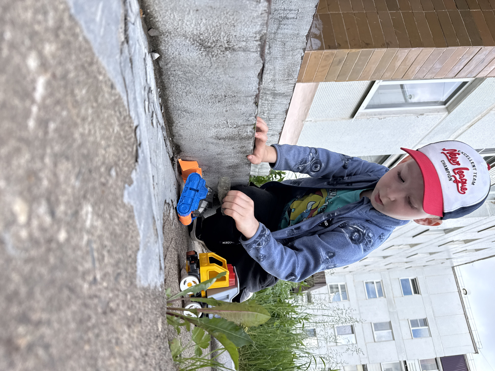 A young boy is captured outdoors, squatting or crouching down, engrossed in playing with two toy trucks near a concrete ledge or wall. The image is rotated 90 degrees clockwise, presenting the scene horizontally.
The boy wears a red and white baseball cap with the text "EXCELLENT TEAM", "New League", and "CHAMPION" visible on it. He is dressed in a blue zipped jacket, a light green t-shirt with a graphic, black pants with "#CROCUS" printed in white on the left leg, and black sneakers. His left hand is resting on the textured concrete ledge, while his right hand is near a yellow and grey toy dump truck. Another orange and blue toy truck is positioned further along the ledge to his left.
The setting appears to be an urban or suburban outdoor area, likely during the daytime under an overcast sky, as suggested by the soft, diffused lighting and lack of strong shadows. Sparse green grass and weeds grow near the concrete, adding a touch of nature to the otherwise built environment. In the background, out of focus, are multi-story residential buildings, one with brown tiled sections and windows, and another lighter-colored building with numerous windows. The boy's expression is calm and focused, indicating he is deeply absorbed in his play.
