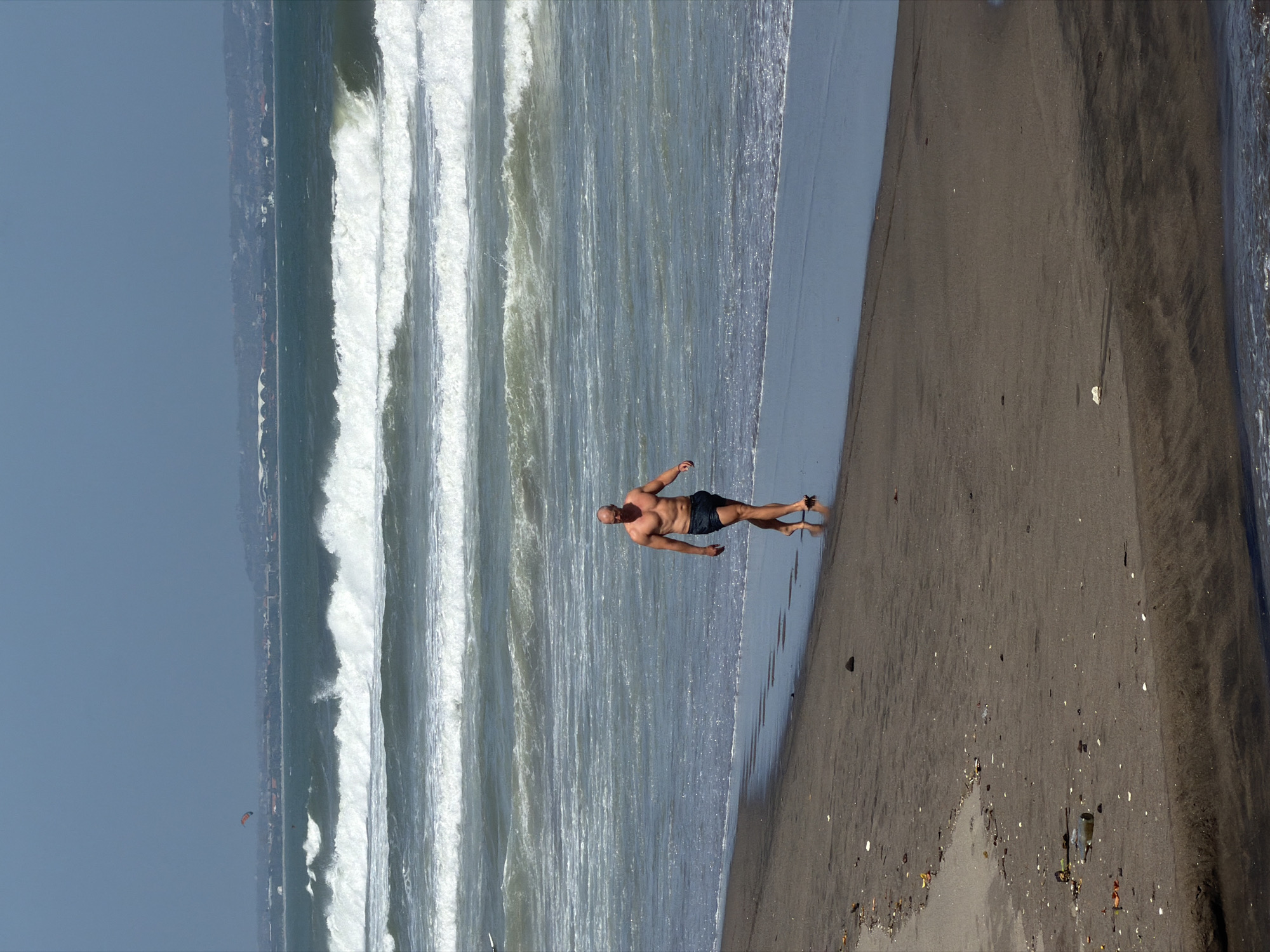 A bald, muscular man walks along a sandy beach toward the right of the frame. He is wearing dark swim trunks and is bare-chested. The man is walking on wet sand near the shoreline, with the ocean to his left. The ocean has large waves crashing with white foam. In the distance, a hazy coastline with buildings can be seen. The sky is a clear, bright blue. The sand on the beach is dark and appears to have some debris scattered on it, including small shells and pieces of vegetation. The scene is likely the coast of Bali, Indonesia, given the appearance of the waves and the general landscape.