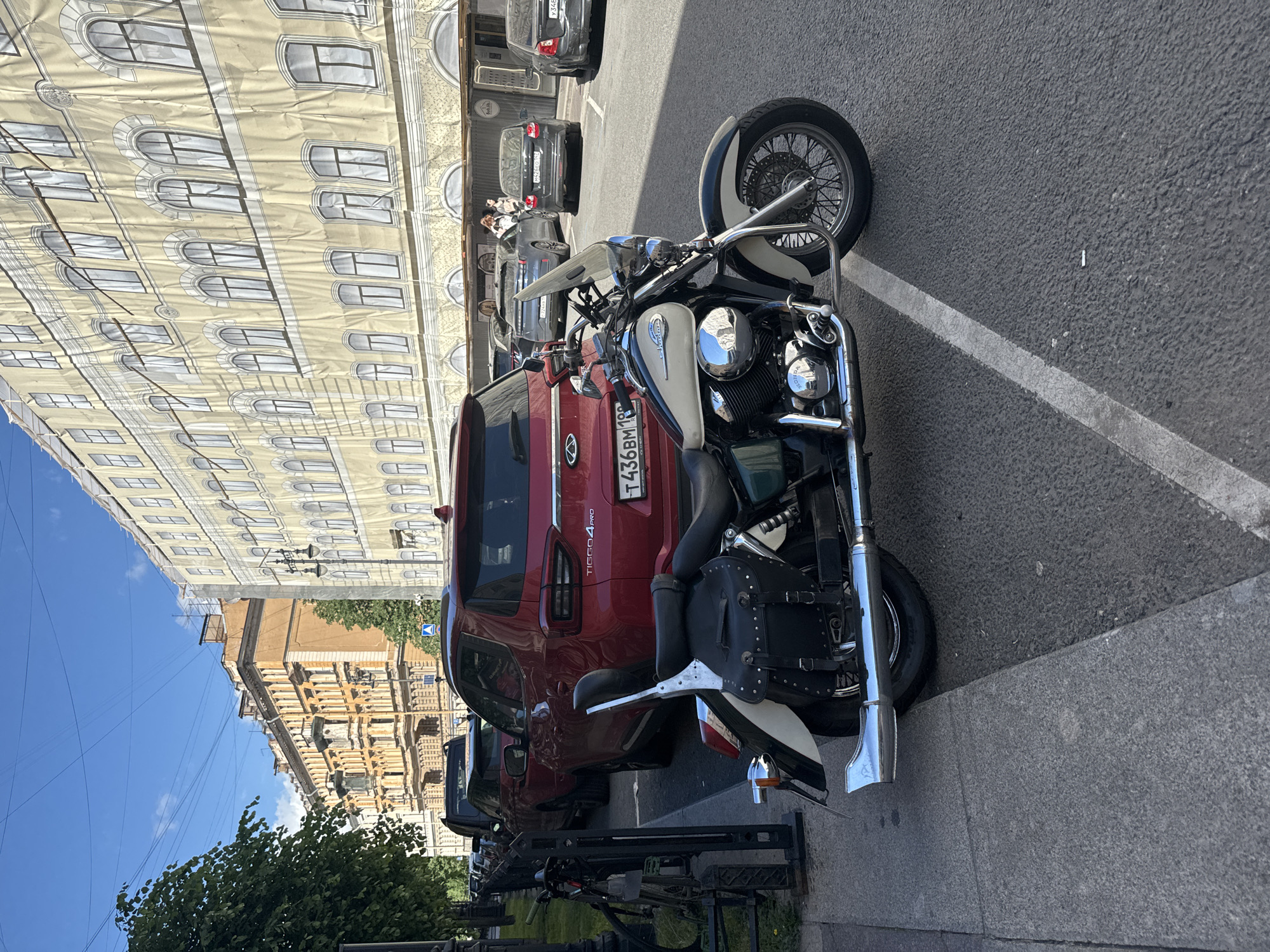 A street scene in Saint Petersburg, Russia, features a parked motorcycle and several cars. The motorcycle, a cruiser-style bike with a white and black paint scheme and chrome accents, is parked on a paved surface next to a red SUV. The SUV, identified by the license plate "T436BM 198", is parked behind the motorcycle.
The background shows buildings with ornate architecture, typical of Saint Petersburg. One building on the left is covered in a large banner or tarp printed with a facade design, suggesting ongoing construction or renovation. Another building to the right displays traditional European architectural elements.
The time appears to be daytime, indicated by the bright sunlight casting shadows. The weather seems clear and sunny. A few individuals are visible in the background, near the cars, but their actions are not discernible. The overall scene suggests a typical urban environment on a pleasant day, with vehicles parked along the street.