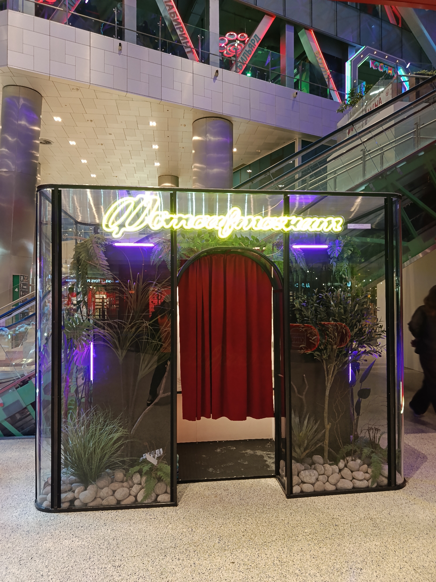 The image shows a photo booth in a shopping mall. The booth has a black frame and glass walls, inside which are potted plants and river rocks, illuminated with purple and green neon lights. A red curtain covers the entrance. Above the booth, neon lettering spells out "Фотобудочки" in a cursive style. The floor is a speckled beige, and the background includes a high ceiling with a white grid, as well as escalators. The mall appears to be in Moscow, Russia, based on the Cyrillic signage visible in the background. A blurry figure of a person in dark clothing is visible in the right edge of the image.
