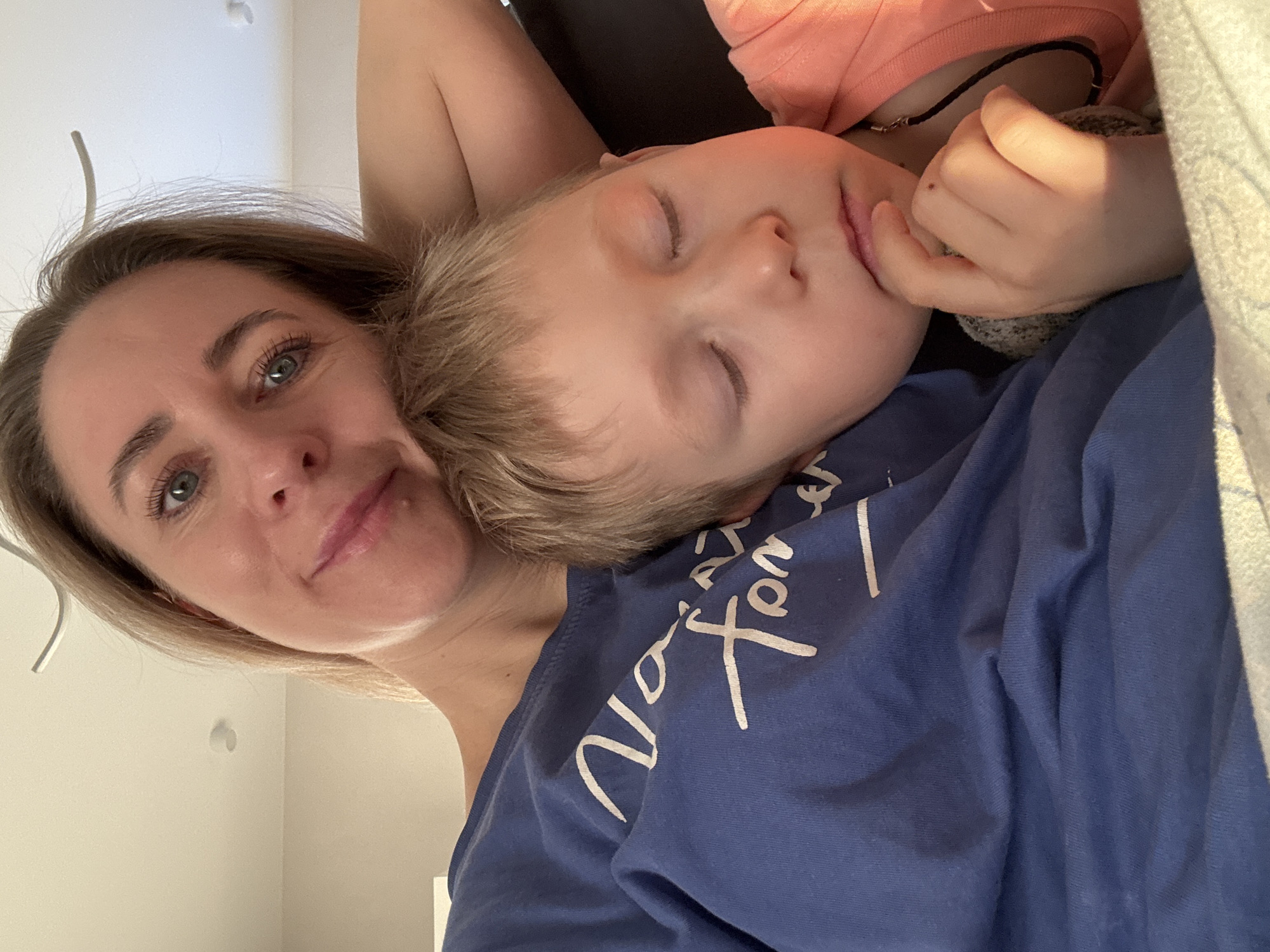 A woman with shoulder-length blonde hair and blue eyes is pictured in a close-up selfie, likely taken indoors given the visible walls and ceiling. She has a slight smile. Lying on her chest is a young boy with blonde hair and closed eyes, indicating he may be sleeping. The boy has another person next to him in a pink shirt with a black necklace. The woman is wearing a blue shirt with white writing on it. The location of Chaikovsky, Russia, cannot be determined from the image.
