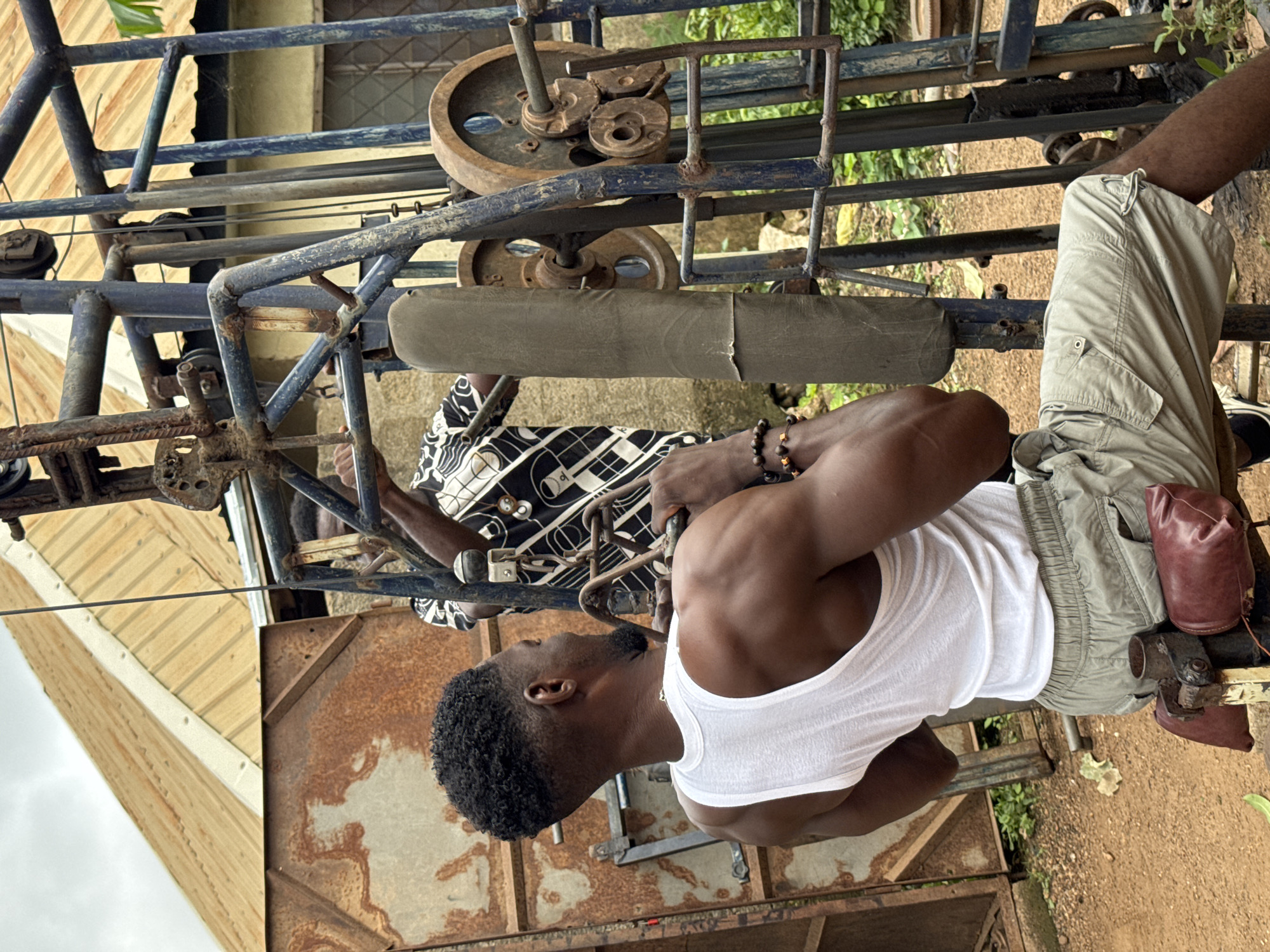 In Akwanga, Nigeria, a man in a white tank top and khaki shorts is performing a lat pulldown exercise on a weathered, rustic weight machine. The machine, constructed from blue metal pipes and rusty components, features weight plates and a padded pull bar. He is seated and actively engaged in the exercise, his back muscles prominently displayed. Behind him, another person in a black and white patterned shirt stands near the machine, observing or assisting. The background reveals a rustic, outdoor environment with corrugated metal structures, dirt ground, and green vegetation, suggesting a local gym or a personal outdoor workout space. The overall scene depicts a moment of physical exertion and training in a functional, somewhat dilapidated setting.