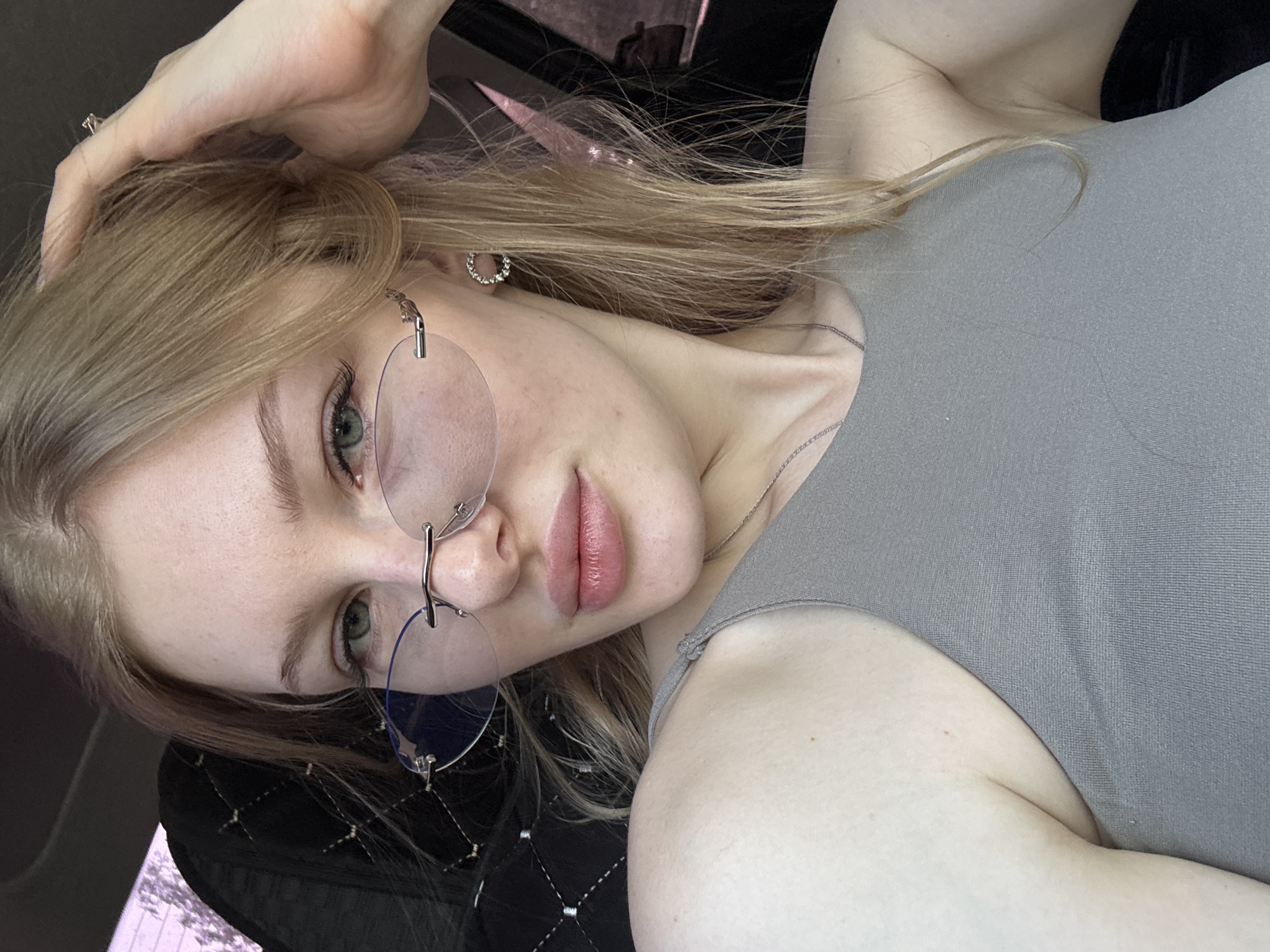 A fair-skinned woman with light blonde hair poses in a tilted headshot. She is wearing rimless, round glasses with silver frames. Her skin appears to be clear, and her lips are a pink shade. She has a silver necklace. She is wearing a gray top. The background appears to be the interior of a car. The exact location in Perm, Russia, cannot be determined from the provided image.