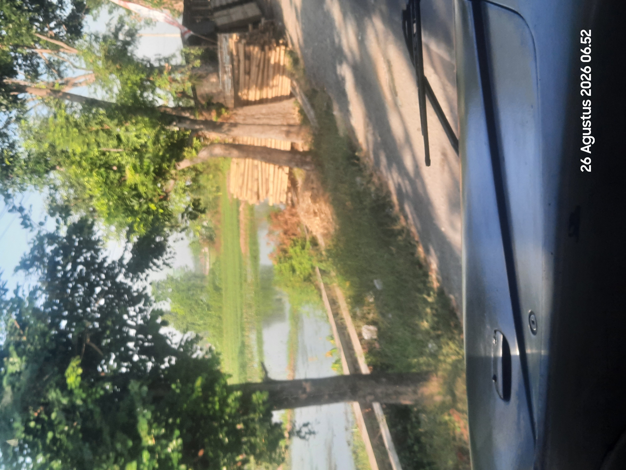 The image captures a view from inside a vehicle, looking out onto a rural scene. In the foreground, the dashboard and windshield wipers of the vehicle are visible. The middle ground features a dirt road, trees, and a body of water that appears to be a pond or flooded field. Stacked lumber is piled on the right side of the road. In the background, there are more trees and a hint of a building. The sky is bright and clear. A timestamp at the bottom right of the image reads "26 Agustus 2026 06.52". The scene suggests a location in Sukokerto, Indonesia, given the date format and the rural setting. There are no people or discernible actions depicted.