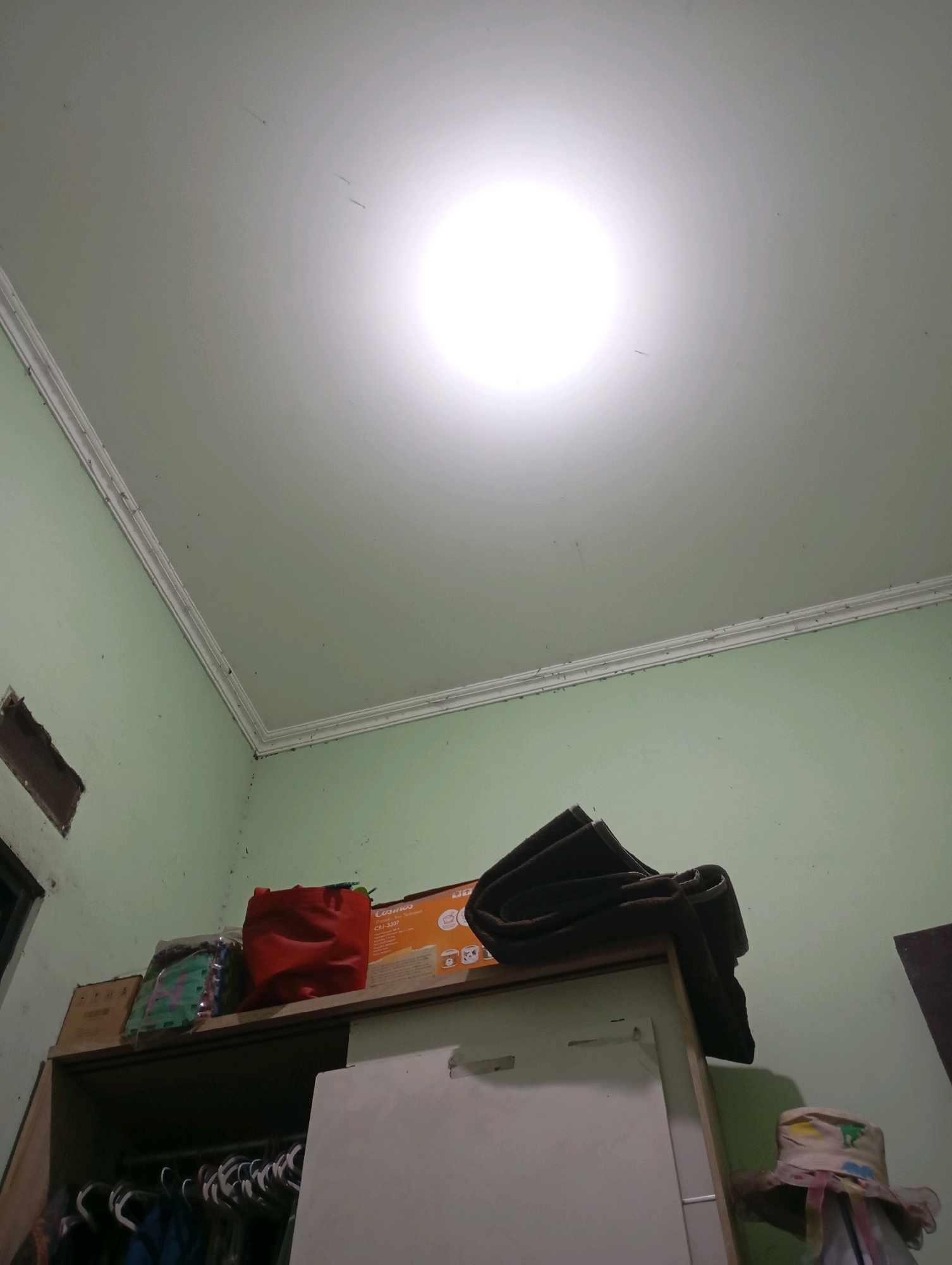 The image shows the interior of a room with light green walls and a white ceiling. A bright light source is visible on the ceiling. In the lower part of the image, there is a tall wooden cabinet. On top of the cabinet, there are several items: a red bag, a box with text that appears to be in Indonesian, some folded fabric items, and a small hat-like object with colorful decorations. Below the cabinet top, clothes are hanging on a rod. The overall impression is of a cluttered storage area within a residential setting. Based on the context of Babelan, Indonesia, this scene likely depicts a domestic storage space.