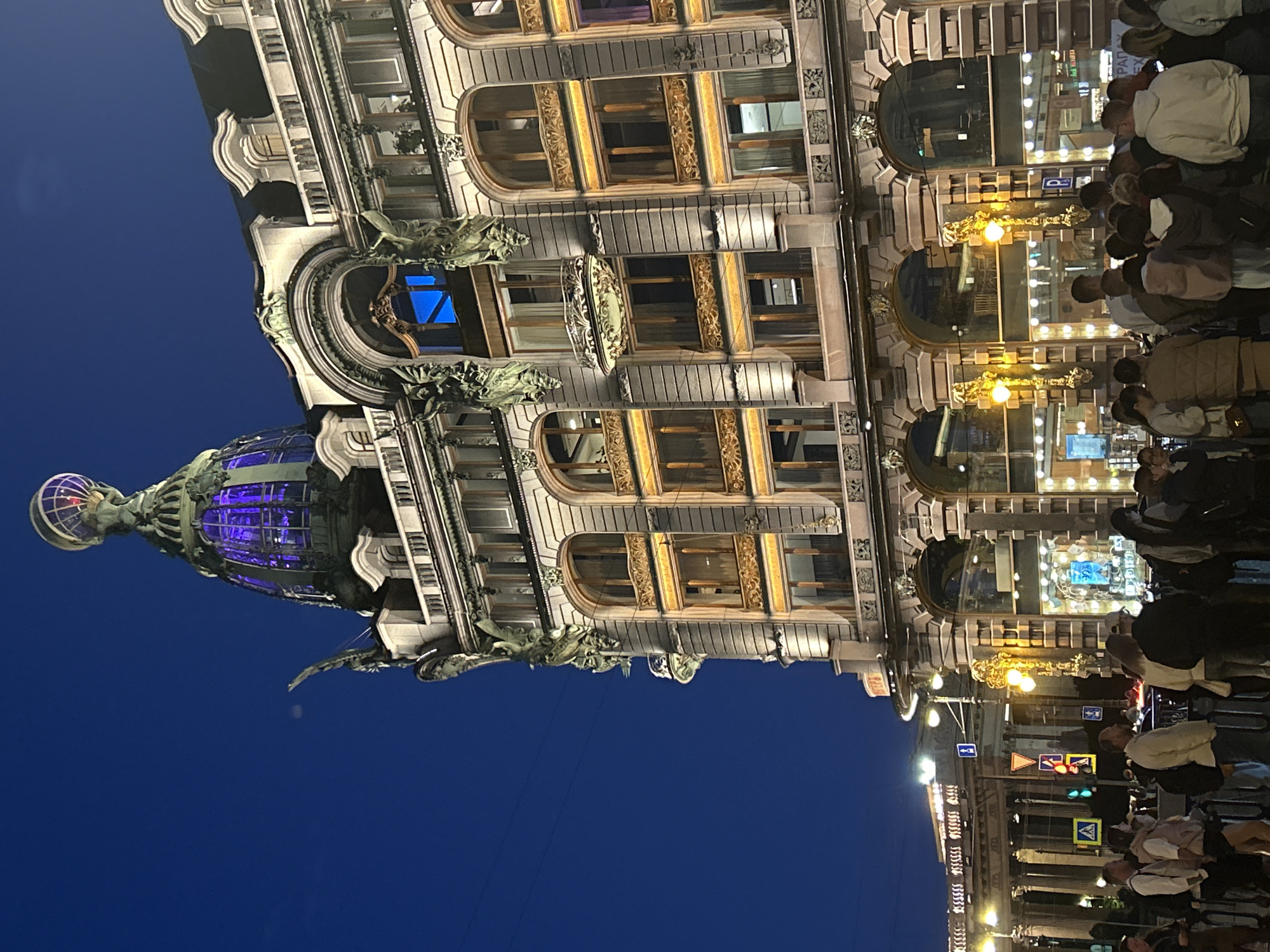 The image shows the Singer House in St. Petersburg, Russia, at twilight. The building's ornate facade, with its detailed sculptures and arched windows, is illuminated by warm light, contrasting with the deep blue of the night sky. A prominent glass-domed cupola, topped with a globe sculpture, sits atop the building, glowing with purple light. A crowd of people is gathered on the sidewalk in front of the building, some looking towards the structure, others engaged in conversation. Streetlights cast a yellow glow, and traffic signals are visible in the background. The overall scene captures the grandeur of the historic architecture under the evening sky.