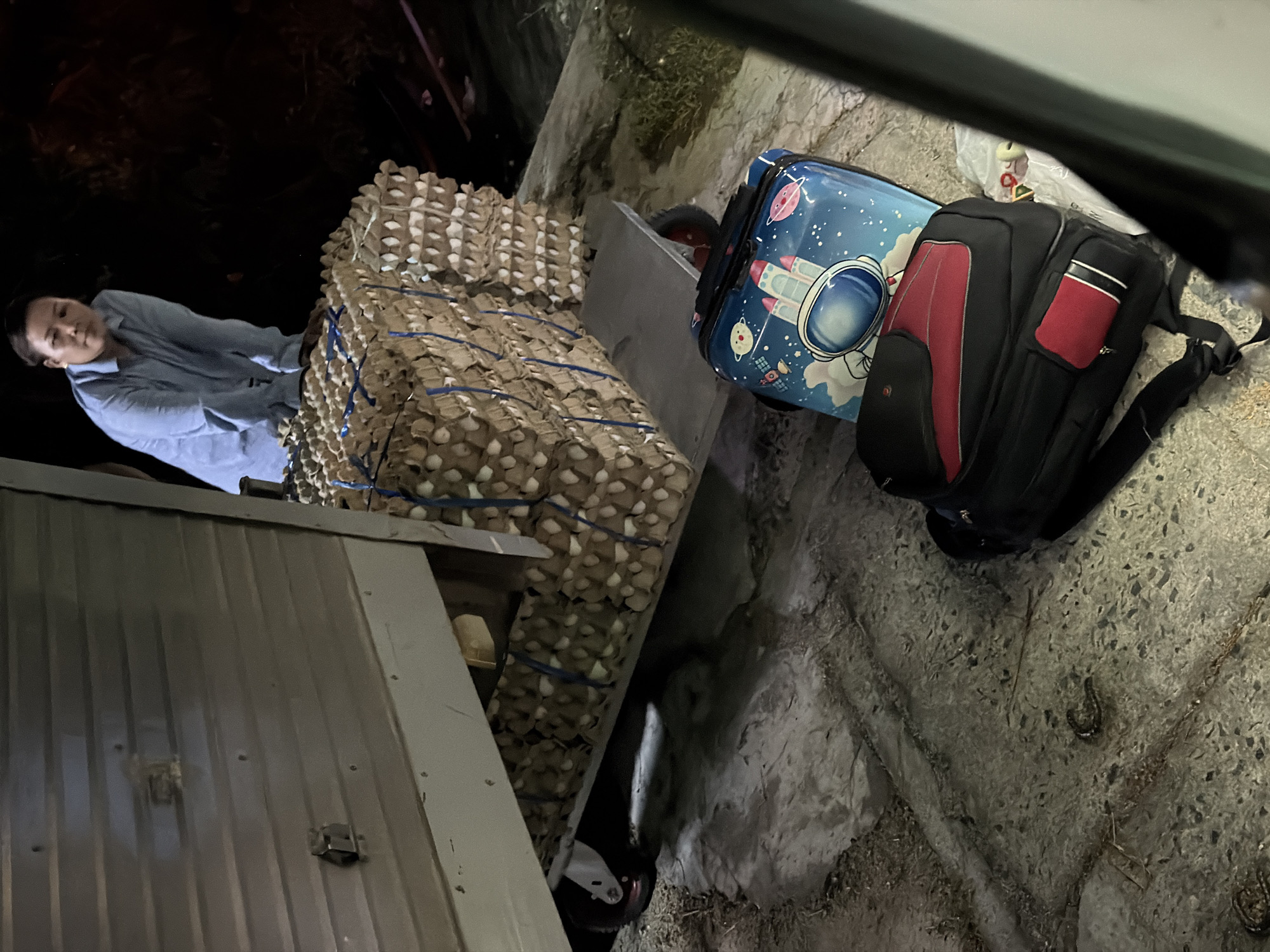 A woman in a light blue collared shirt is leaning over a large stack of egg cartons. The egg cartons are secured with blue straps and are stacked on a wheeled cart. The cart is positioned beneath the open rear of a light-colored trailer, suggesting loading or unloading activity. In the foreground, two pieces of luggage are visible on the ground: a black and red backpack and a small blue suitcase adorned with an astronaut and space-themed designs. The scene appears to be outdoors, with a concrete or paved surface and some vegetation visible in the background. The lighting suggests it is nighttime or dusk. The location is identified as Đại Lãnh, Vietnam.