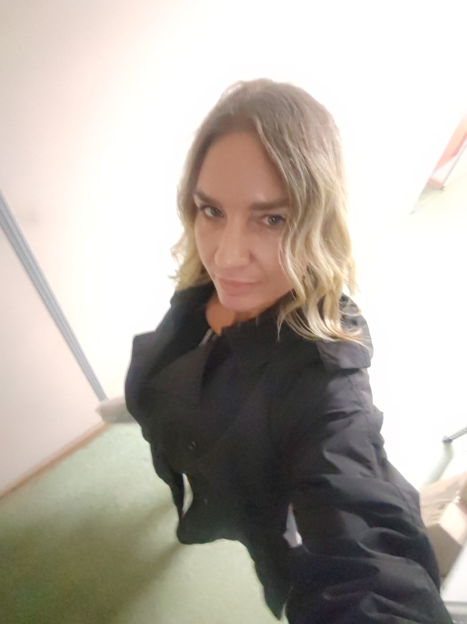 A woman with blonde wavy hair is indoors, wearing a black coat. The background is a blurred mix of white walls, green carpet, and what appears to be office furniture. The city and specific location are unknown.