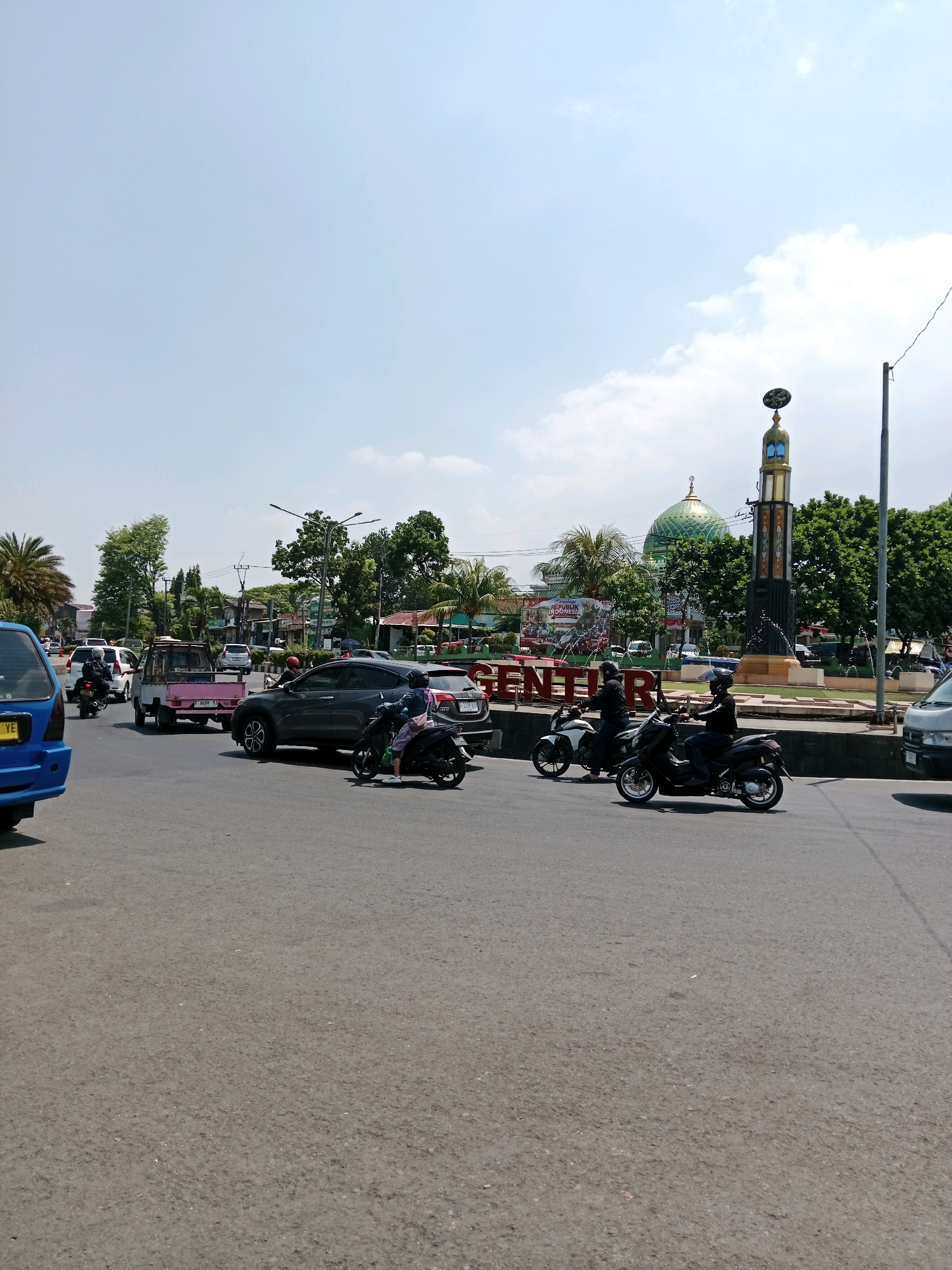 The image captures a daytime street scene in Cianjur, Indonesia, likely near a prominent landmark. In the foreground, asphalt pavement shows tire marks and a few scattered debris. Several vehicles are present, including a blue car on the left, a silver SUV in the middle, and a light-colored truck behind it. A white pickup truck with a pink side panel is also visible. Two individuals on motorcycles are riding on the road.
In the background, a large monument with a golden dome, likely a mosque, stands tall, surrounded by trees and other buildings. The sky is bright blue with scattered white clouds. The overall environment appears to be a busy urban intersection.
