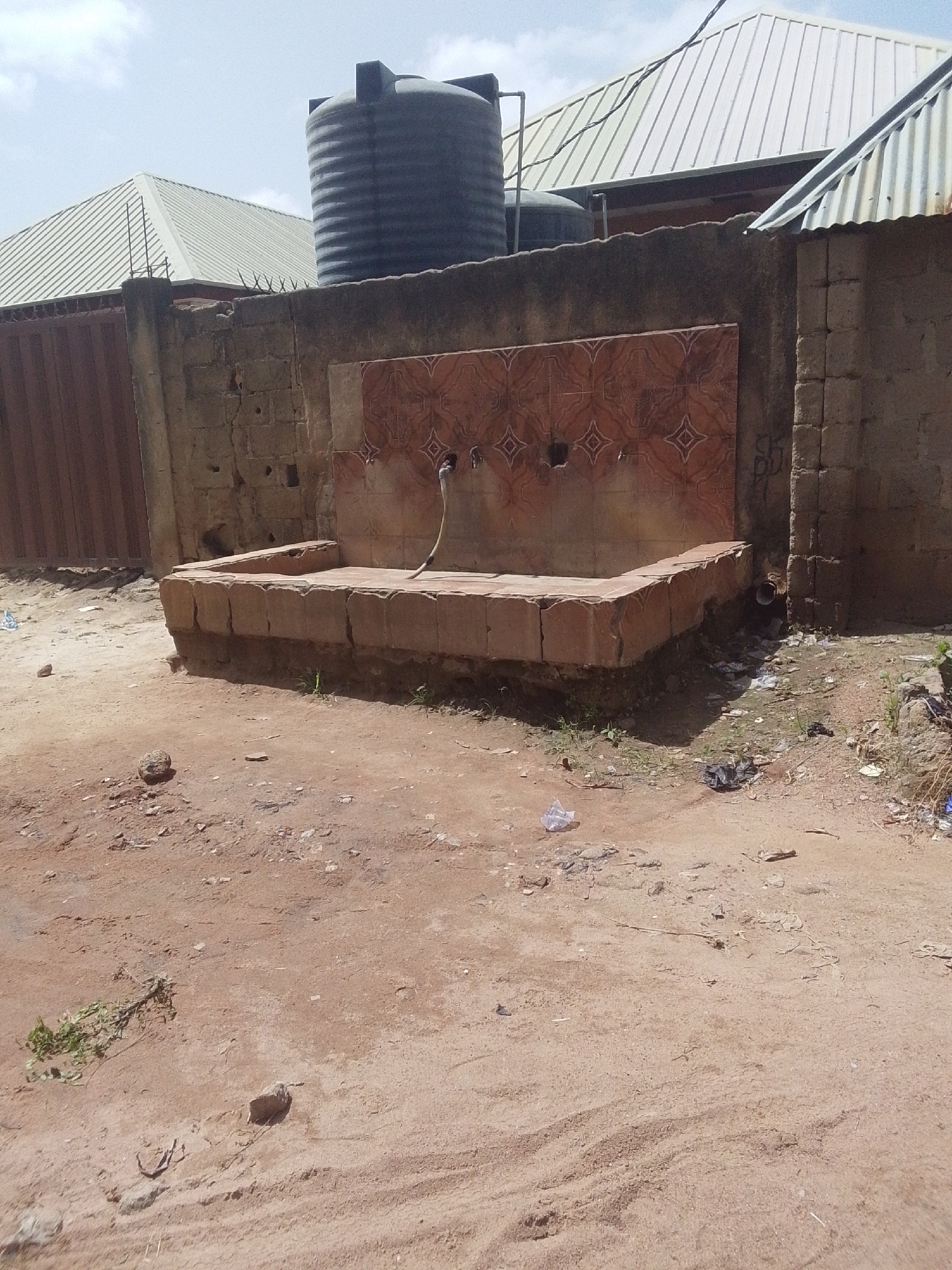 This is an eye-level, medium shot of a public water fountain in Jalingo, Nigeria. The fountain is made of stone and features three spigots. The wall behind the fountain is decorated with patterned tiles. In the background, two water tanks are visible on top of a building with a corrugated metal roof. The ground in front of the fountain is dirt and littered with debris. The sky is bright and overcast.