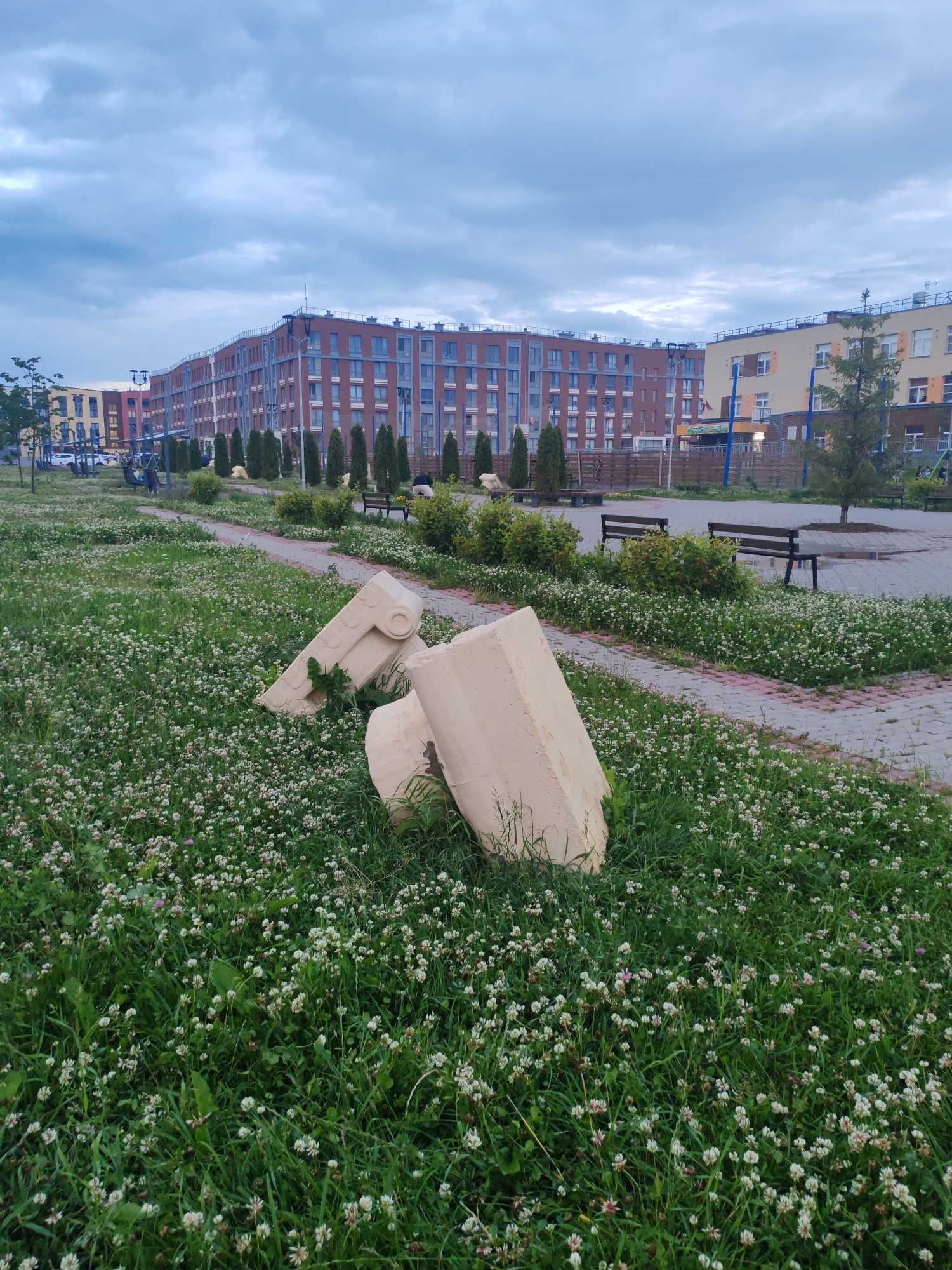 In the city of Shushary, Russia, a ground-level view captures a residential complex with modern brick buildings under a cloudy sky. In the foreground, a patch of green grass dotted with white clover flowers is disrupted by several large, beige, abstract sculptural elements, possibly made of concrete, lying on their sides. A paved walkway with a pink border winds through the grassy area, leading towards the buildings. Benches are strategically placed along the walkway, some with people sitting on them. Trees and shrubs are interspersed throughout the landscape, contributing to the urban park-like setting. The overall scene suggests a public space within a residential area, possibly a courtyard or a small park.