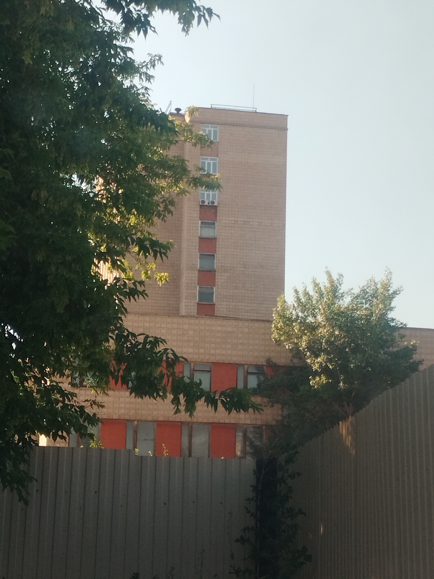 A tall, brick building with multiple floors and many windows is partially visible behind a metal fence and trees. The building's facade is light brown with horizontal bands of orange and grey sections on the lower floors. The windows are rectangular, and some have air conditioning units installed. Trees with green leaves are in the foreground, obscuring parts of the building. The sky is a clear, pale blue. The scene appears to be outdoors in an urban environment.