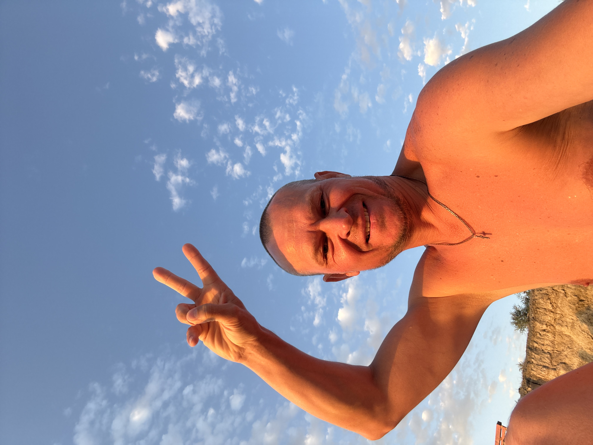 A shirtless man with a shaved head and a goatee smiles at the camera, holding up his right hand in a peace sign against a bright blue sky with scattered clouds. He is wearing a silver chain with a cross pendant. The man's skin is lit by a warm, golden light, suggesting it is either sunrise or sunset. In the background, a rocky cliff face and some sparse vegetation are visible. The overall scene suggests an outdoor setting, possibly near the coast or a natural elevation. The precise location in Zharbulak, Kazakhstan, cannot be determined from the visual information alone.