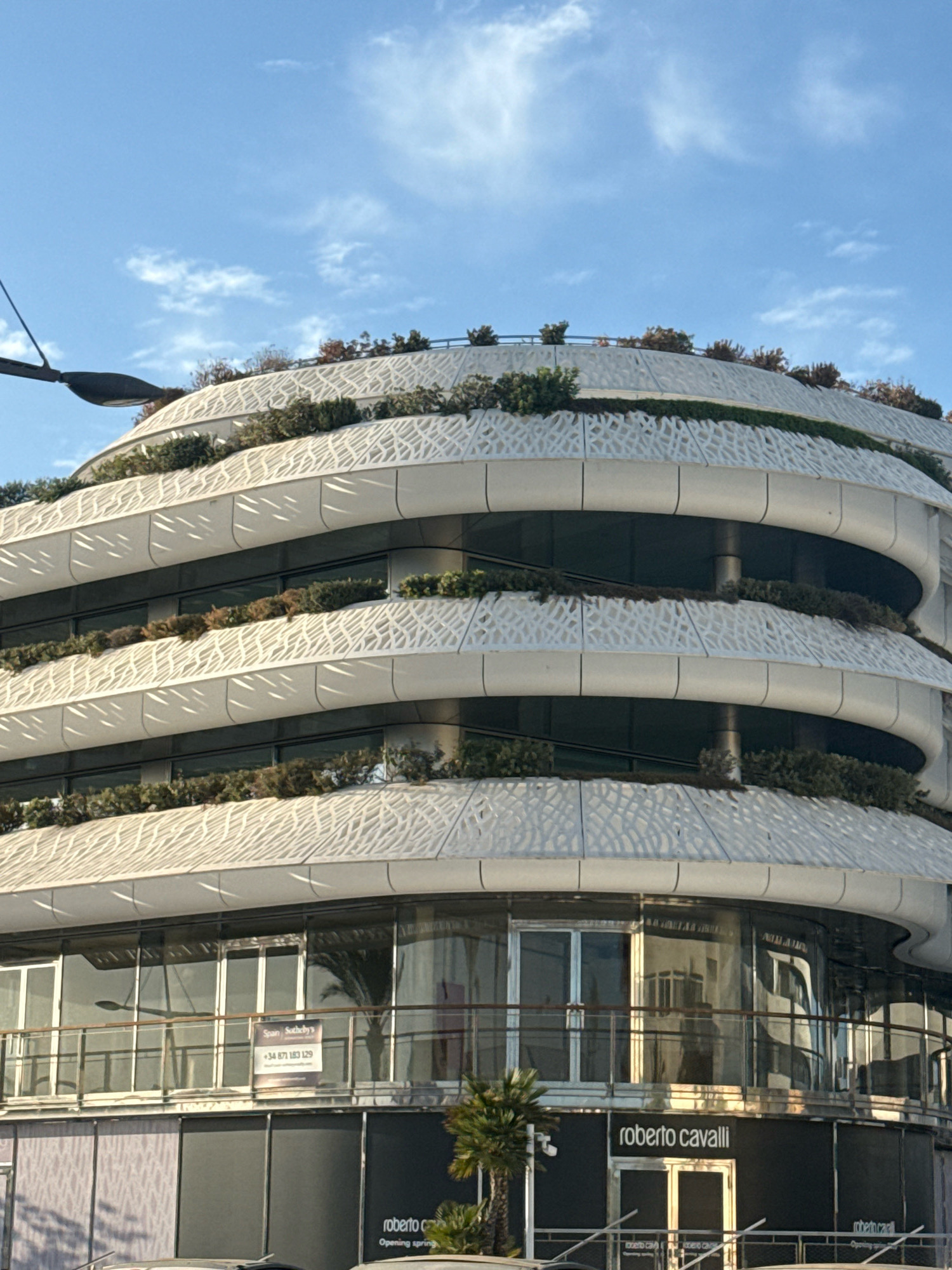 The image captures the exterior of a modern, multi-tiered building with a curved facade, located in Ibiza, Spain. The architecture features white, patterned panels with rounded edges, creating a layered effect. Greenery, likely plants and bushes, is integrated into the design, appearing on terraces and along the edges of each tier. The building has large glass windows and doors on the lower levels, reflecting the surrounding environment. A sign for "Spain Sotheby's" with a phone number is visible on one of the windows. Below the glass facade, dark panels display "roberto cavalli" branding, indicating a retail establishment, with text suggesting an "Opening spring". A palm tree is situated in front of the building. The sky is blue with scattered white clouds, and a street light or part of an antenna is visible in the upper left corner.