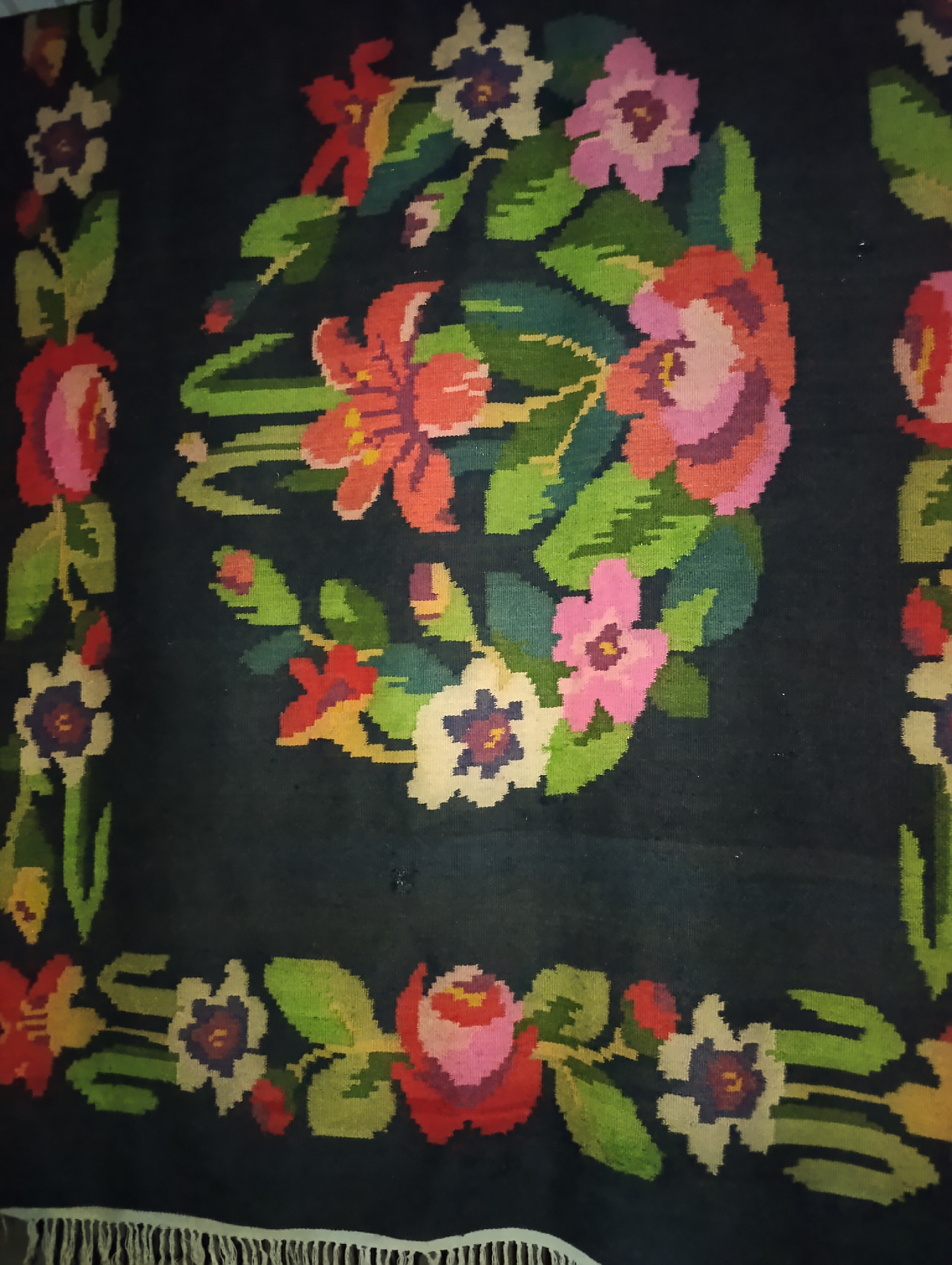 A dark, possibly navy blue, rug with a floral pattern is shown in the image. The rug features a central circular arrangement of flowers and leaves, with a border of floral motifs running along the sides and bottom. The flowers are depicted in various colors, including pink, red, white, and purple, with green leaves. The bottom of the rug has a fringe. The image appears to have been taken indoors, potentially in a home in Roghi, Moldova, as suggested by the context. There are no people or animals visible in the image.