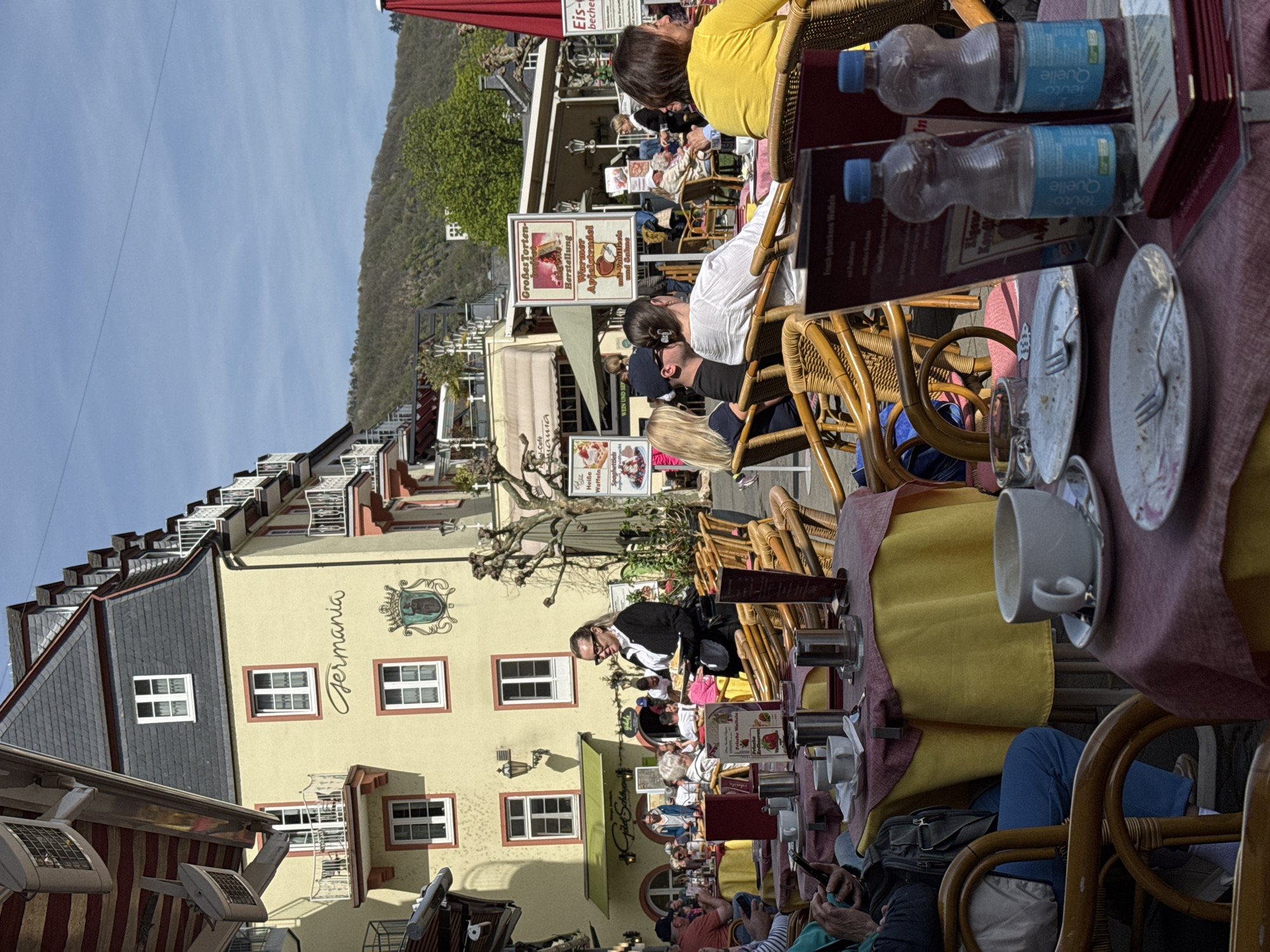 The image depicts a bustling outdoor café in Cochem, Germany, during daylight. People are seated at tables, eating and drinking. Wicker chairs and tables with red and yellow tablecloths fill the foreground. A waitress is seen attending to customers. "Germania" is written on the facade of a building. In the background, other buildings and trees are visible under a blue sky. Signage advertises "Heiße Waffeln" and "Großes Torten." Water bottles and menus are on the tables.