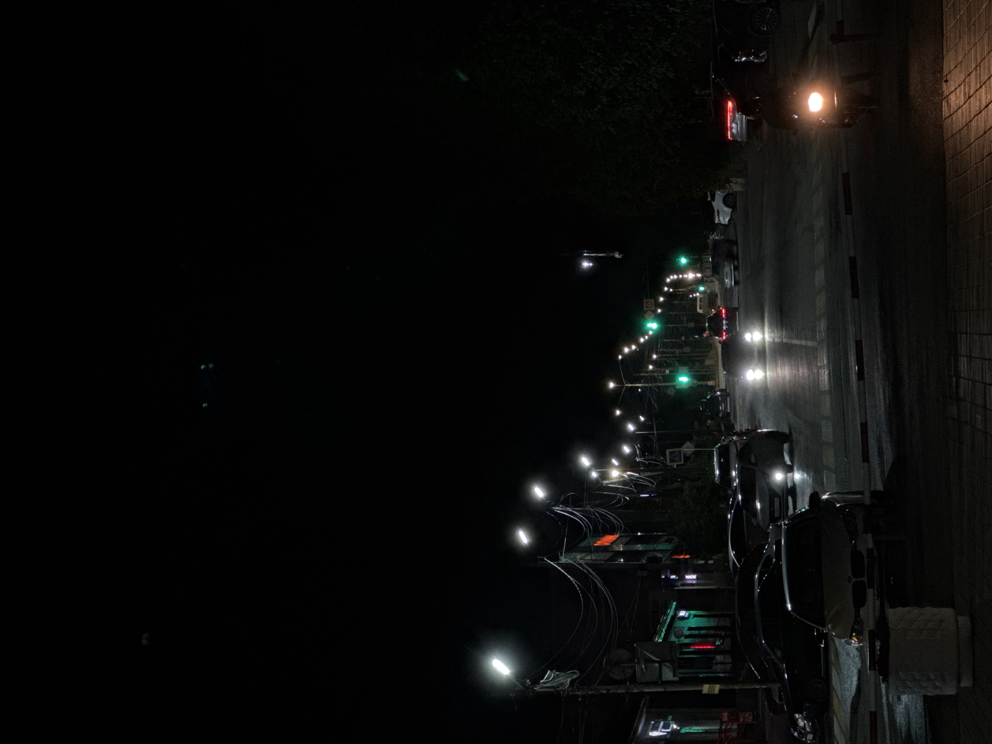 The media file depicts a night scene in Mikhaylovsk, Russia, likely on a street. The image is oriented vertically.
The primary subjects are various vehicles, including cars and possibly a motorcycle, parked or moving along a wet road. There are streetlights and traffic signals illuminating the scene. Some buildings and trees are visible along the side of the road, creating a city or town atmosphere. The ground appears wet, suggesting recent rain or damp conditions.
The activity appears to be a typical urban night, with streetlights casting glows and vehicle lights reflecting on the wet pavement. There is no clear indication of specific events or people prominently featured, as the focus is on the nocturnal urban environment.
Notable details include the bright lights of street lamps and traffic signals, the reflections on the wet surface, and the silhouettes of vehicles. The time of day is clearly night, as indicated by the darkness and artificial lighting. No specific weather conditions are definitively discernible beyond the wetness of the ground. Location cues are limited to the context provided that it is Mikhaylovsk, Russia. No text is visible in the image. The overall mood is calm and quiet, characteristic of a late-night urban setting.