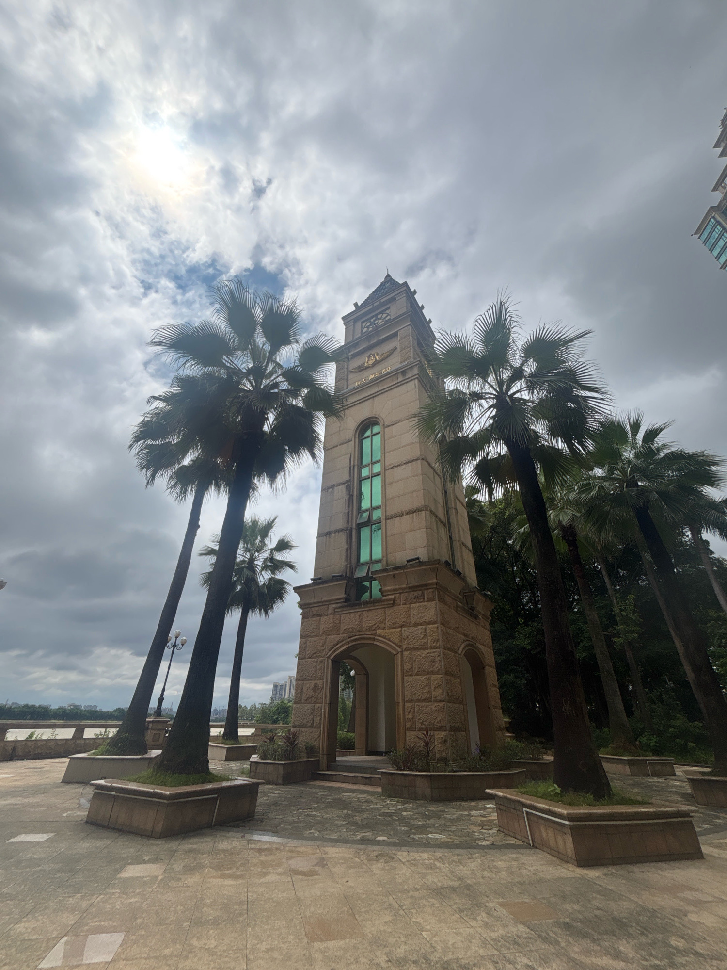 The image shows a tall, stone clock tower with a green-tinted window, surrounded by palm trees on a cloudy day. The clock tower has a classical architectural style with arched entrances at its base. The ground is paved with stone tiles. In the background, a body of water, likely a river, is visible, with buildings on the opposite bank. The overall scene suggests a public park or waterfront area in a city, possibly with a slightly humid climate given the presence of palm trees. The image was taken from a low angle, looking up towards the clock tower and the sky. The location is Nanhai District, China.