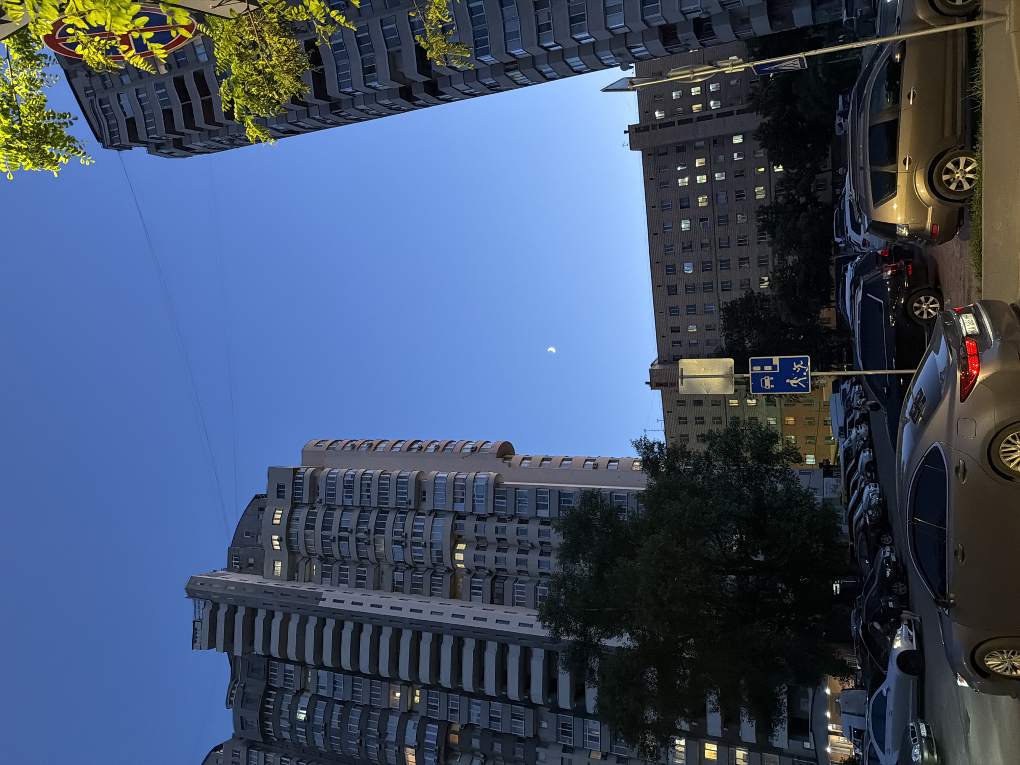The image depicts a street scene at dusk in Kyiv, Ukraine, featuring several multi-story apartment buildings and a parking lot filled with cars. The sky is a deep blue, with a sliver of the moon visible.
On the left, a tall, curved apartment building with numerous balconies dominates the foreground. Its facade is light gray, and many windows are illuminated, suggesting occupancy. To the right of this building, a shorter, older apartment building with a brick facade is visible, also with lit windows.
The right side of the image is occupied by another tall, curved apartment building, this one appearing to be darker gray. Green foliage from trees is encroaching on the upper right portion of this building, and a street sign with a red, white, and blue circular emblem is partially visible.
In the middle ground, a parking lot is filled with various vehicles, including sedans and SUVs. A woman in dark clothing is standing near the hood of a white car, seemingly interacting with it. Several traffic and parking signs are present, including a blue square sign with pictograms of a car, a person walking, and a wheelchair symbol.
The foreground shows the asphalt of the parking lot and a small strip of grass and curb. The overall impression is a typical residential area in a large city during the evening hours.