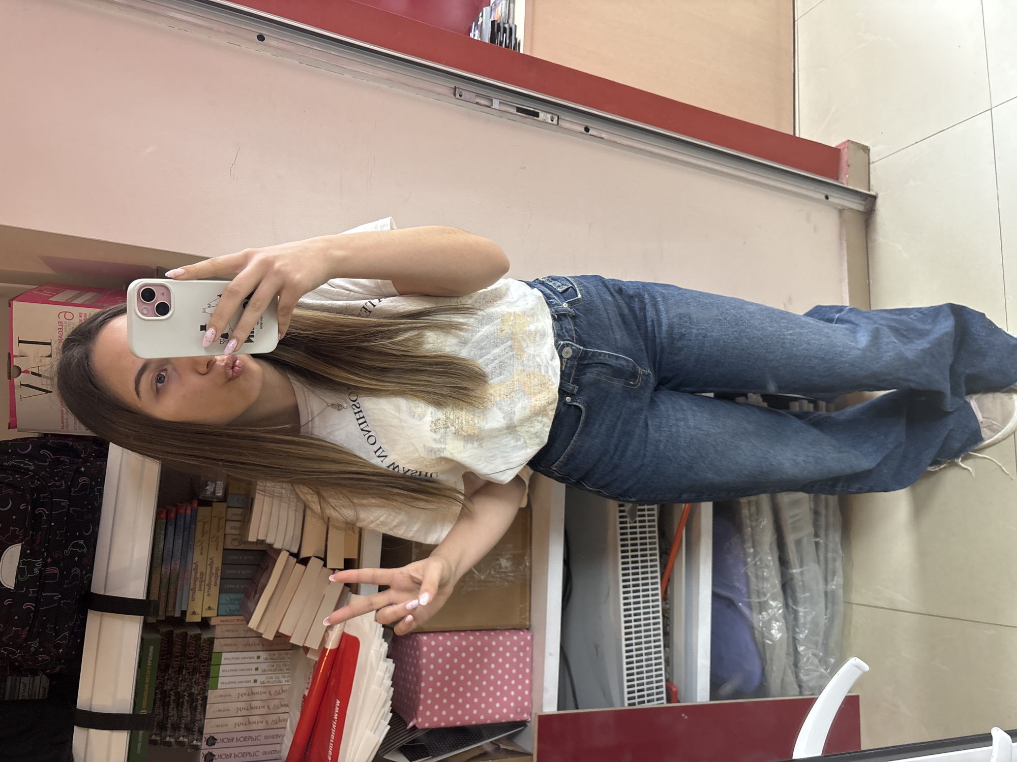 A young woman is taking a selfie in front of a bookshelf filled with books, a backpack, and other items. She is holding a phone with a white case and making a peace sign with her left hand. She is wearing a white t-shirt with writing on it and blue jeans. The background appears to be a store or library setting. The image is rotated. Some text on the bookshelf is Cyrillic, and some text on a red sign reads “www.vivacom.bg.” Given the Vivacom reference, it is likely the location is in Bulgaria. The exact location within the city of Veliko Tarnovo, Bulgaria, cannot be determined from the image alone.