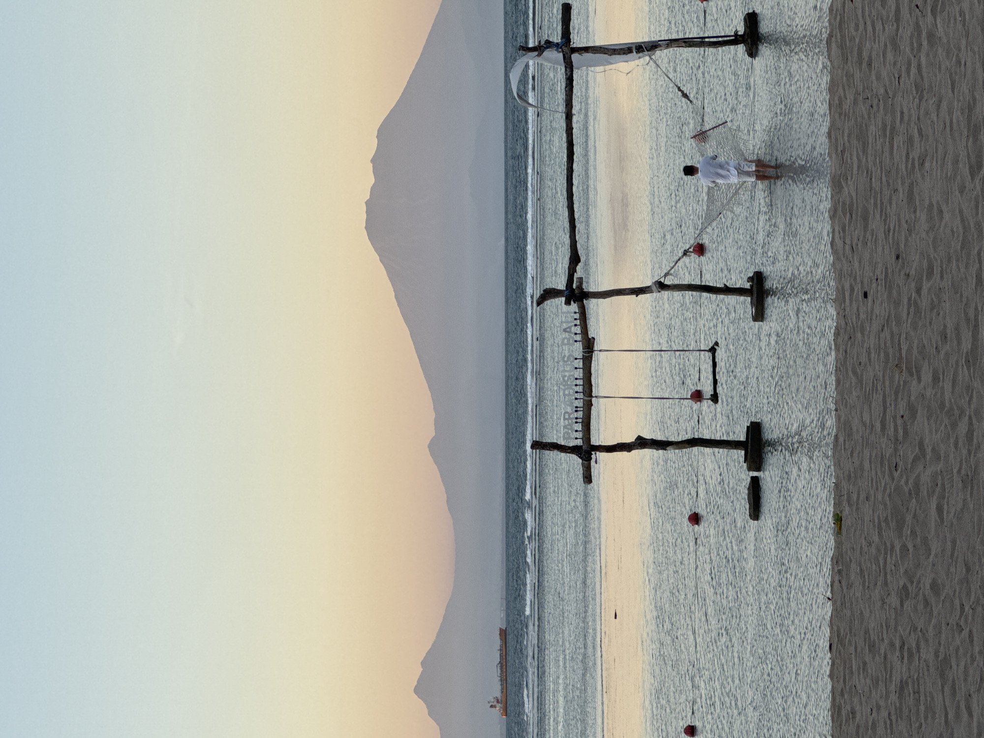 This image depicts a serene coastal scene, possibly at sunset or sunrise, in Kuta Selatan, Indonesia. The right side of the image features a sandy beach. The foreground shows wooden structures with ropes and red buoys anchored in the water. A man, dressed in white, is walking in the water near one of the structures. The sea stretches towards the horizon, with gentle waves. In the background, there is a mountainous landscape against a sky with gradients of light blue, yellow, and orange. The overall atmosphere appears tranquil and scenic.