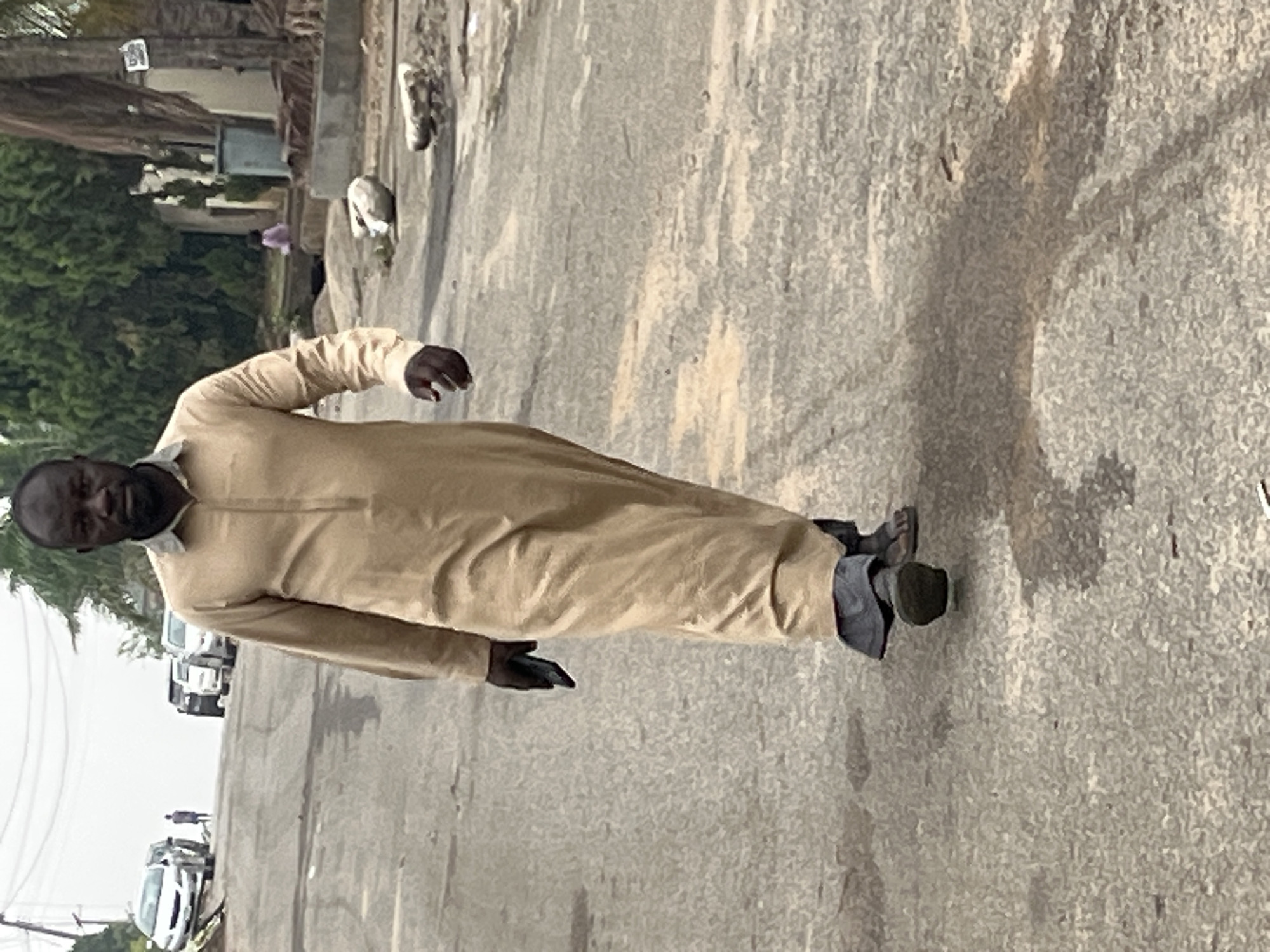 A middle-aged man wearing a traditional tan-colored robe and sandals walks on an asphalt road in Abuja, Nigeria. He is holding a black smartphone in his left hand. Several vehicles, including cars and a truck, are parked on the road behind him. Trees are visible in the background.