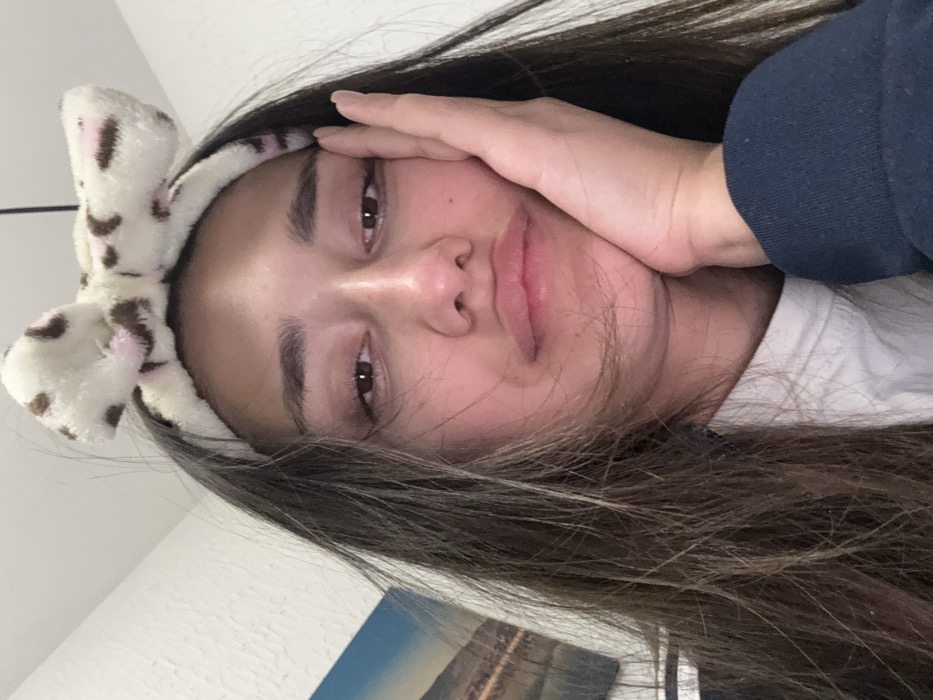 A young woman with long, brown hair poses for a selfie. She wears a fuzzy headband with a bow, decorated with brown and pink spots. Her skin appears smooth and fair. One hand is raised to her forehead, partially obscuring her face, while the other hand is likely supporting her chin. Her lips are slightly pursed, and her gaze is soft. The background is blurry, but glimpses of a white wall and a framed picture with blue hues can be seen.