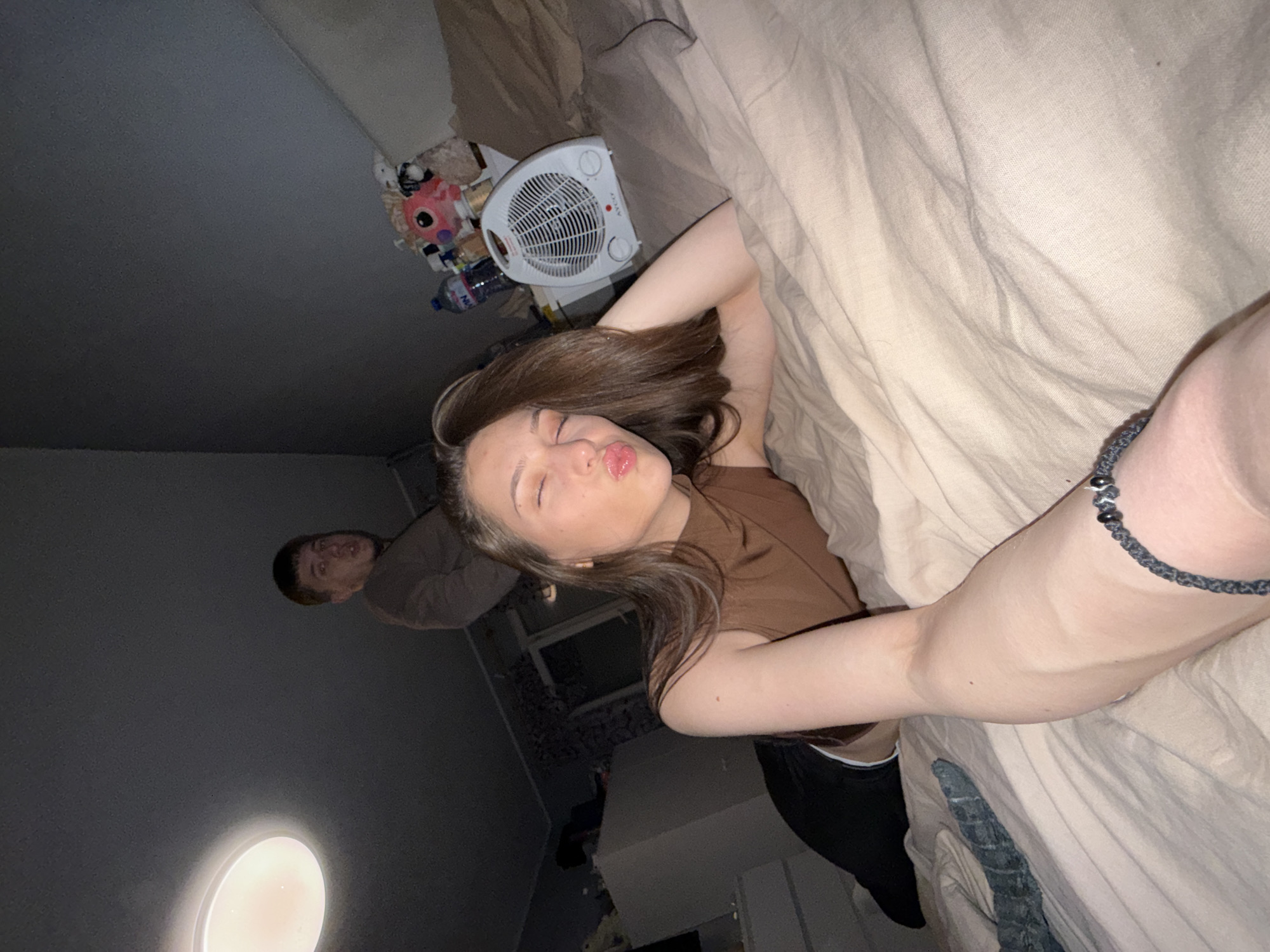 The image depicts an indoor scene, likely a bedroom, with dim lighting suggestive of evening or nighttime. The primary subject is a young woman, positioned diagonally across the frame, appearing to take a selfie or a casual photo. She has long, dark brown hair and is wearing a brown sleeveless top and dark pants. Her eyes are closed, and her lips are playfully pursed in a "kiss" expression. Her right arm, adorned with a dark beaded bracelet, rests on a light-colored duvet or blanket that covers much of the right side of the frame. Her left arm is raised, with her hand behind her head.
In the upper left background, partially visible behind the woman, a young man looks towards the camera with a neutral or slightly smiling expression, wearing a long-sleeved top. The room appears to be casually furnished; a white, circular electric fan or space heater with a red "AYCO" logo is visible on a surface in the upper right, next to plastic bottles (including a water bottle) and several stuffed toys, notably a pink one. A circular light fixture illuminates a portion of the wall in the lower left background. The overall mood conveyed by the woman's pose and expression is playful and relaxed. The setting is consistent with a residential interior.