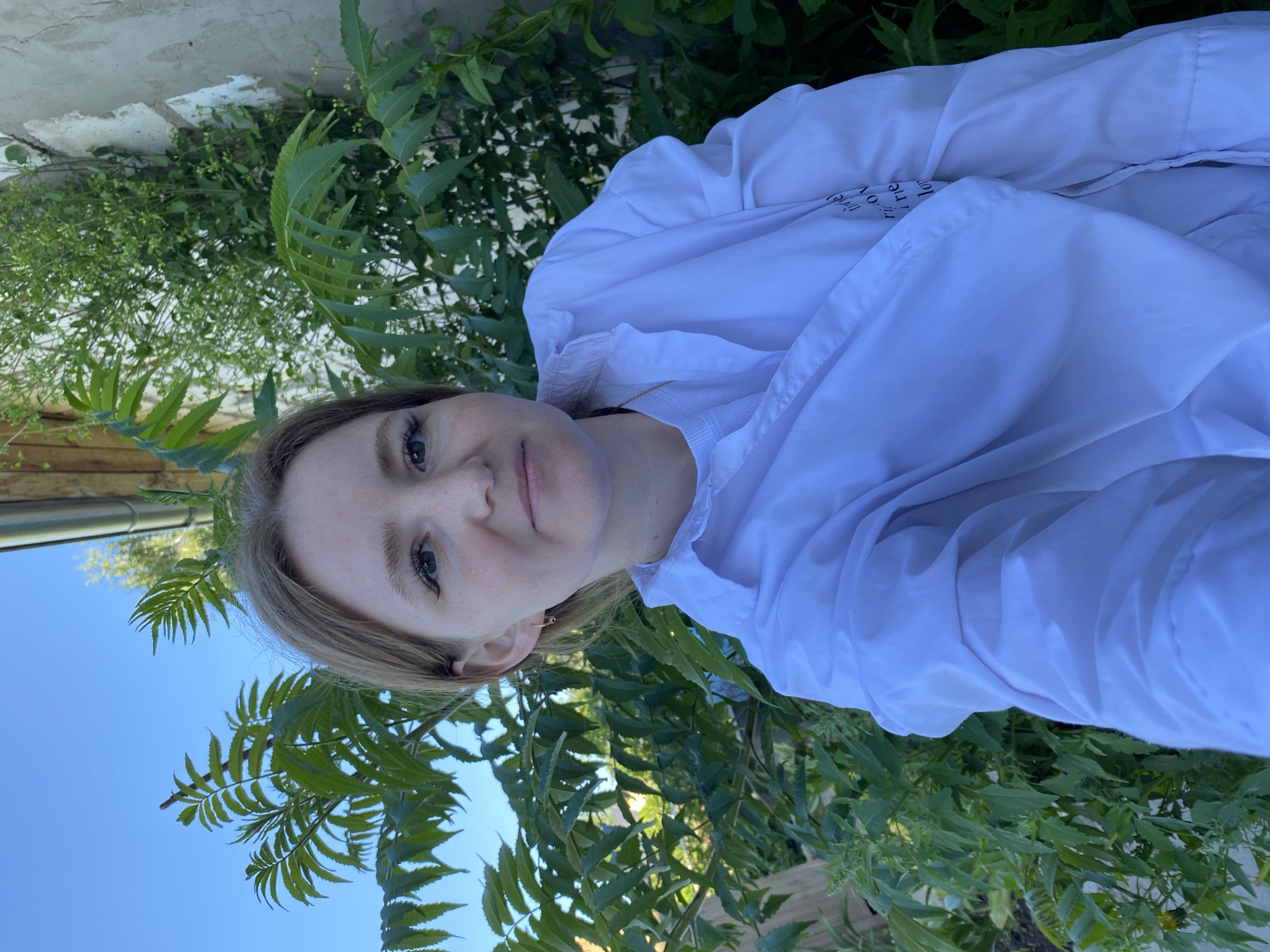 In an outdoor setting, a young Caucasian woman with light brown hair and light skin, exhibiting freckles, poses for a selfie. She is wearing a white collared shirt over a light-colored top and a delicate gold earring in her left ear. Her makeup includes eyeliner and mascara. The background features lush green foliage and a textured wall, with hints of a wooden structure and a gutter visible at the top. The scene appears to be in Chaikovsky, Russia, likely a residential area given the presence of the wall and foliage. The lighting suggests it is daytime, with the sun casting a soft glow. There are no other individuals or discernible actions in the frame.