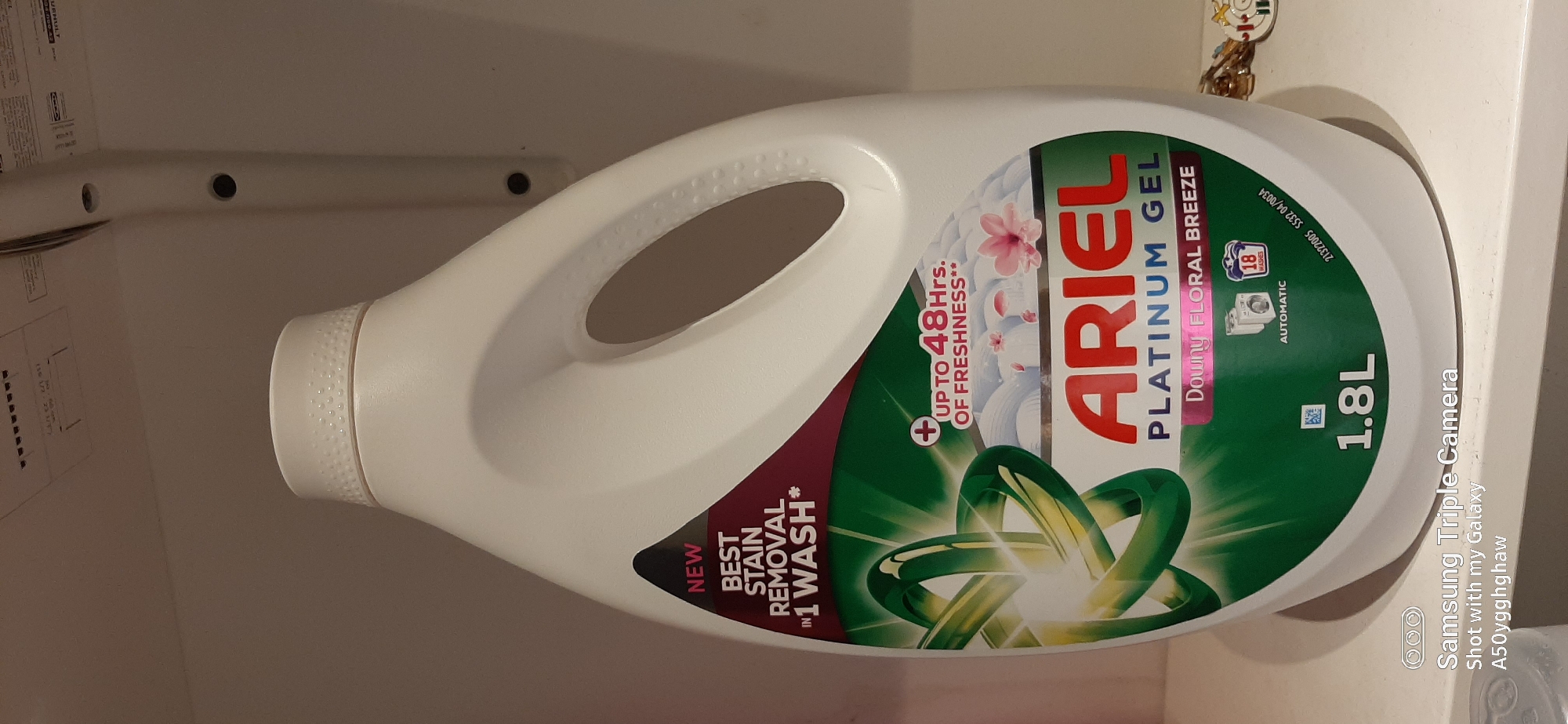 The image shows a white plastic bottle of Ariel Platinum Gel laundry detergent, which is standing on a white shelf. The bottle has a large handle and a white cap. The label on the bottle is primarily green and red, with text and graphics advertising its features. The text includes "NEW BEST STAIN REMOVAL IN 1 WASH*", "UP TO 48 HRS. OF FRESHNESS**", "ARIEL PLATINUM GEL", "Downy FLORAL BREEZE", and "1.8L". There are also icons depicting a washing machine and "18 WASHES". A watermark at the bottom of the image indicates "Samsung Triple Camera Shot with my Galaxy A50yggghghaw". The background appears to be a white wall with some indistinct shelving or wall fixtures. The scene is indoors, and the lighting is artificial. There is no indication of the city of Dubai or the United Arab Emirates in the image.