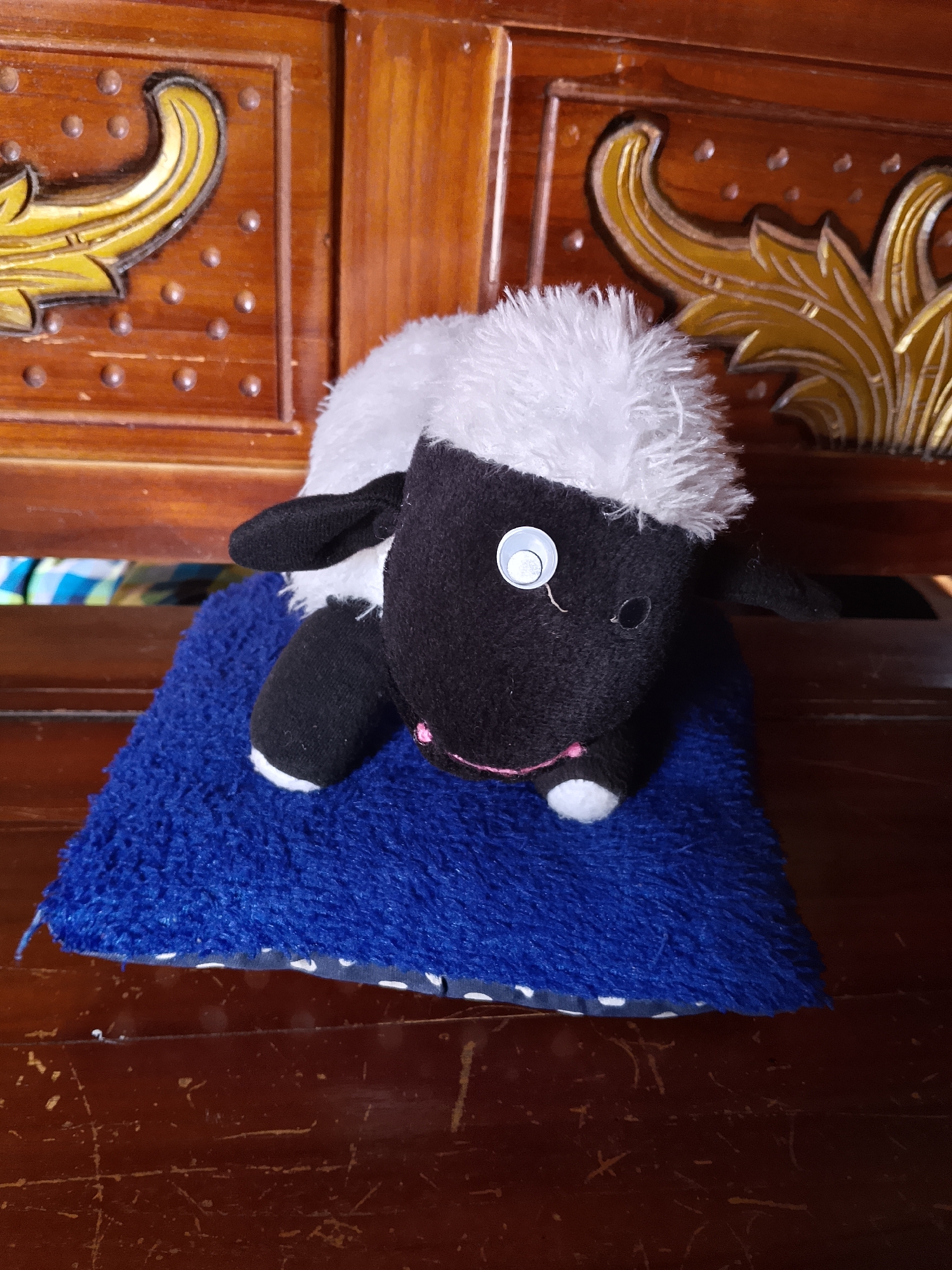 A stuffed toy sheep with black and white fur sits on a blue fluffy mat. The sheep has one googly eye and a pink stitched mouth. It is positioned on the mat, which is placed on a wooden surface. The background features a wooden piece of furniture with ornate golden carvings and dark brown wooden planks. This scene is located in a home in the city of Kasiman, Indonesia.