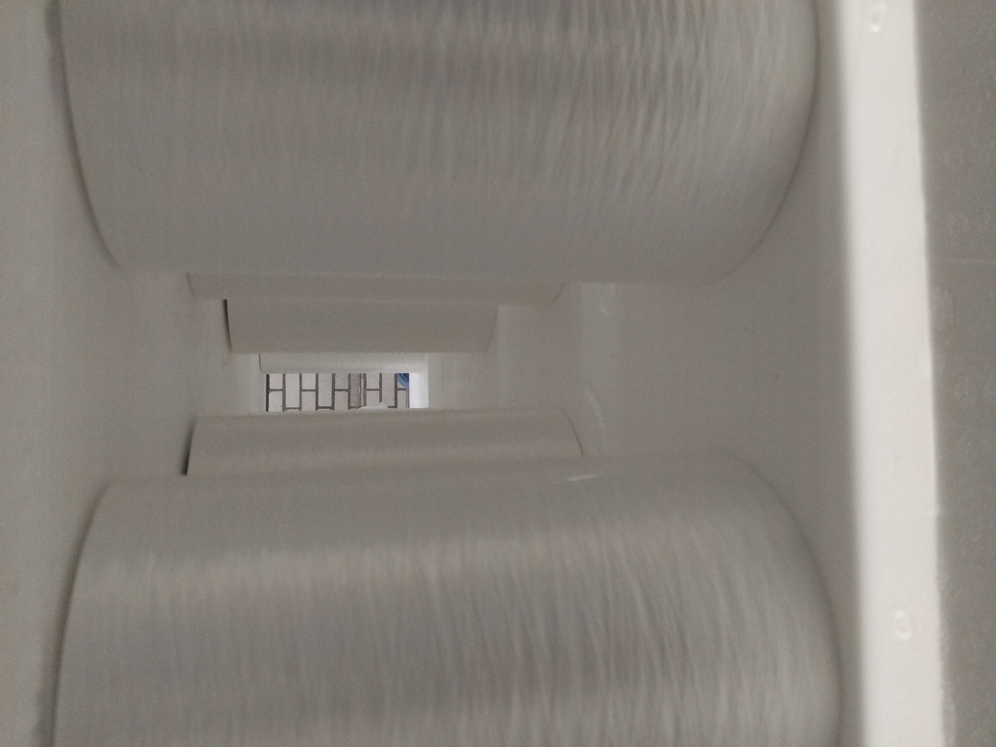 The image shows a perspective view of several large, white, cylindrical spools of material, possibly yarn or fiber, arranged in a row. The spools are tightly wound and have a textured surface. They are positioned within a large, brightly lit, indoor space with a white, textured ceiling and floor. In the background, a section of a brick wall is visible, along with what appears to be a shelf or partition. The overall impression is one of an industrial or storage environment. This image was taken in Deurne, Netherlands, likely within a manufacturing or warehousing facility. There are no people or animals visible in the image.