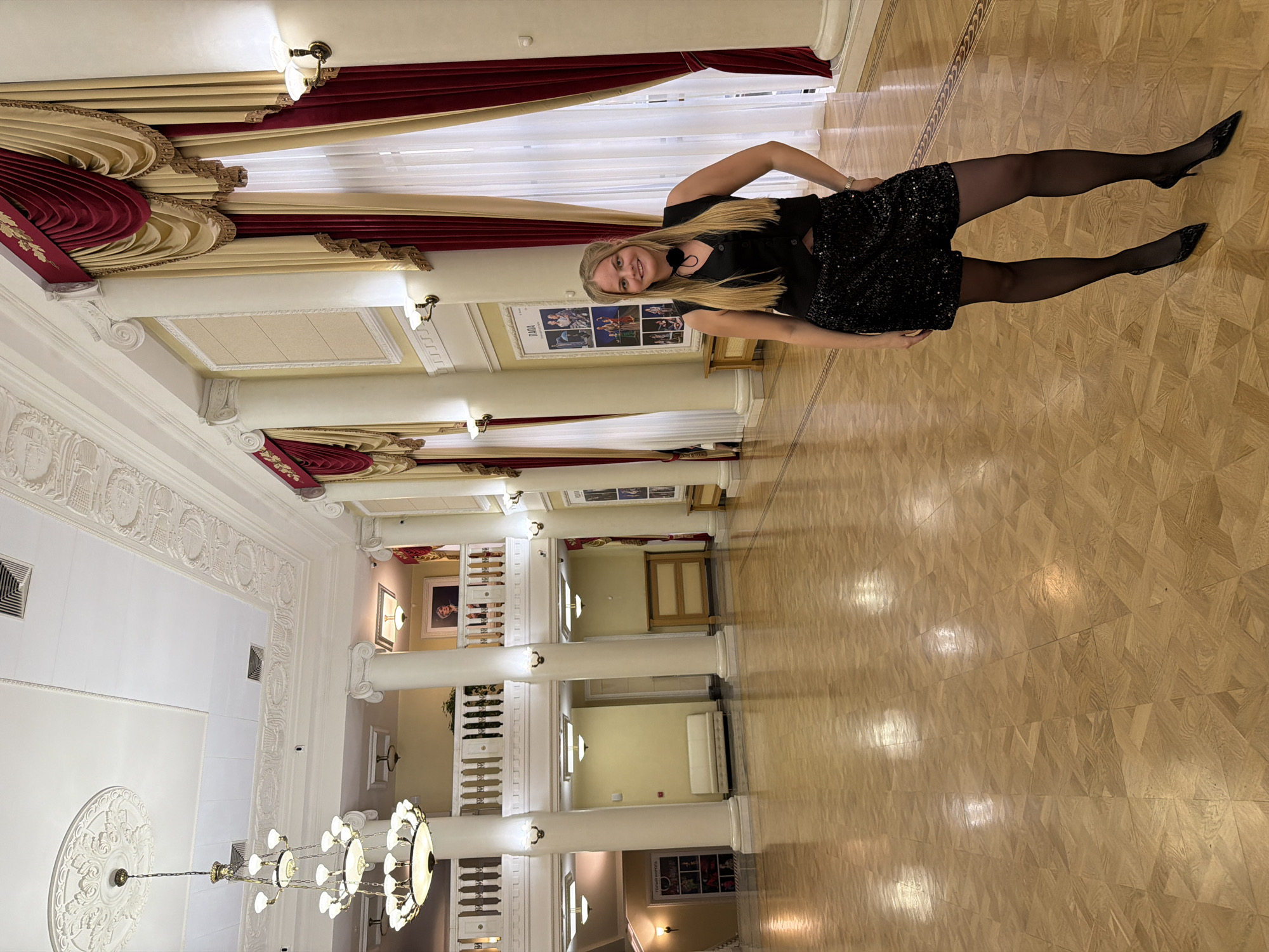 The photograph depicts a woman in a black dress and heels standing in an ornate building. The location is the State National Theater of the Udmurt Republic in Izhevsk, Russia. The woman is blonde and is smiling. The building has a high ceiling with columns, decorative molding, and chandeliers. Red and gold drapes are visible. The floor has a parquet pattern.
