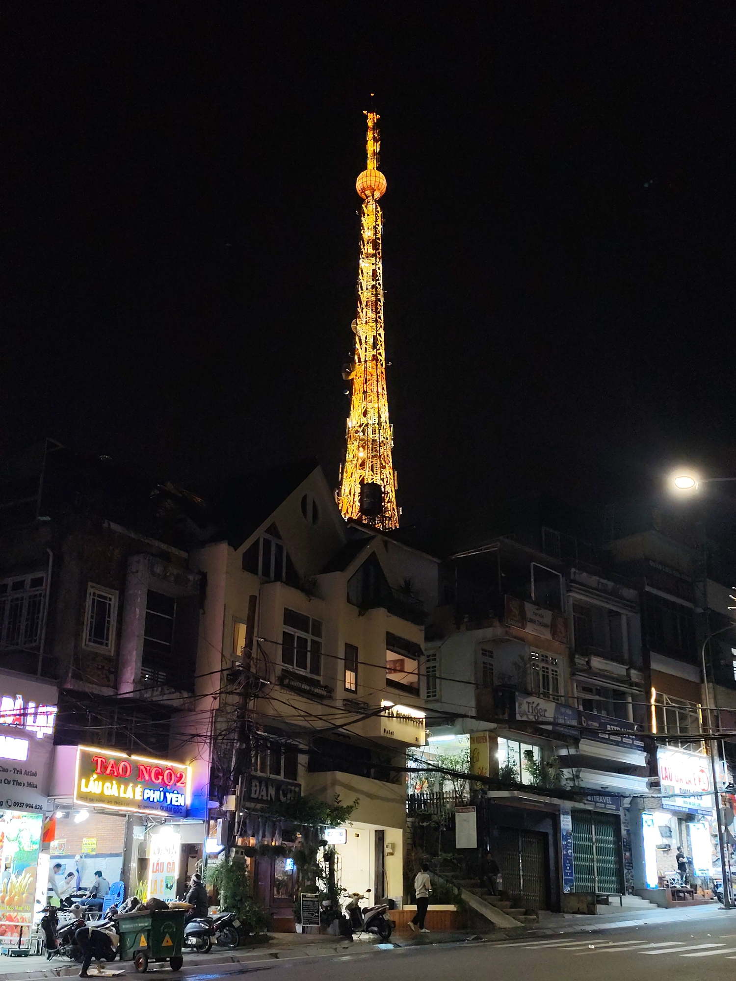 The image captures a vibrant night scene in Da Lat, Vietnam, featuring a tall, brightly illuminated tower rising prominently above a street lined with multi-story buildings. The tower, resembling a communication or observation structure, glows with a warm, golden light against the dark night sky.
At street level, various businesses are open, indicated by their illuminated signs and storefronts. Several people are visible on the sidewalks, some sitting at tables, others walking or standing, contributing to a lively atmosphere. Numerous motorcycles and motorbikes are parked along the curb, a common sight in Vietnamese cities. Overhead, a dense network of electrical wires and cables crisscrosses between the buildings. A green utility cart or garbage bin is also seen on the sidewalk.
Visible text on signs includes "TAO NGO2" and "LẨU GÀ LÁ É PHÚ YÊN" (Chicken hotpot with E leaves, Phu Yen style), indicating a restaurant. Other partial texts like "ĐAN CERT," "QUẦN GỐC," "Tiger," and "TRAVEL" suggest other establishments such as a shop or a travel agency. Further text "Cao-Trà Atiso Cơ The Mùa" and "Bếp Nam Mỹ" suggest a tea shop and a restaurant specializing in Southern American cuisine. The presence of these businesses and the general activity portray a bustling urban environment after dark.