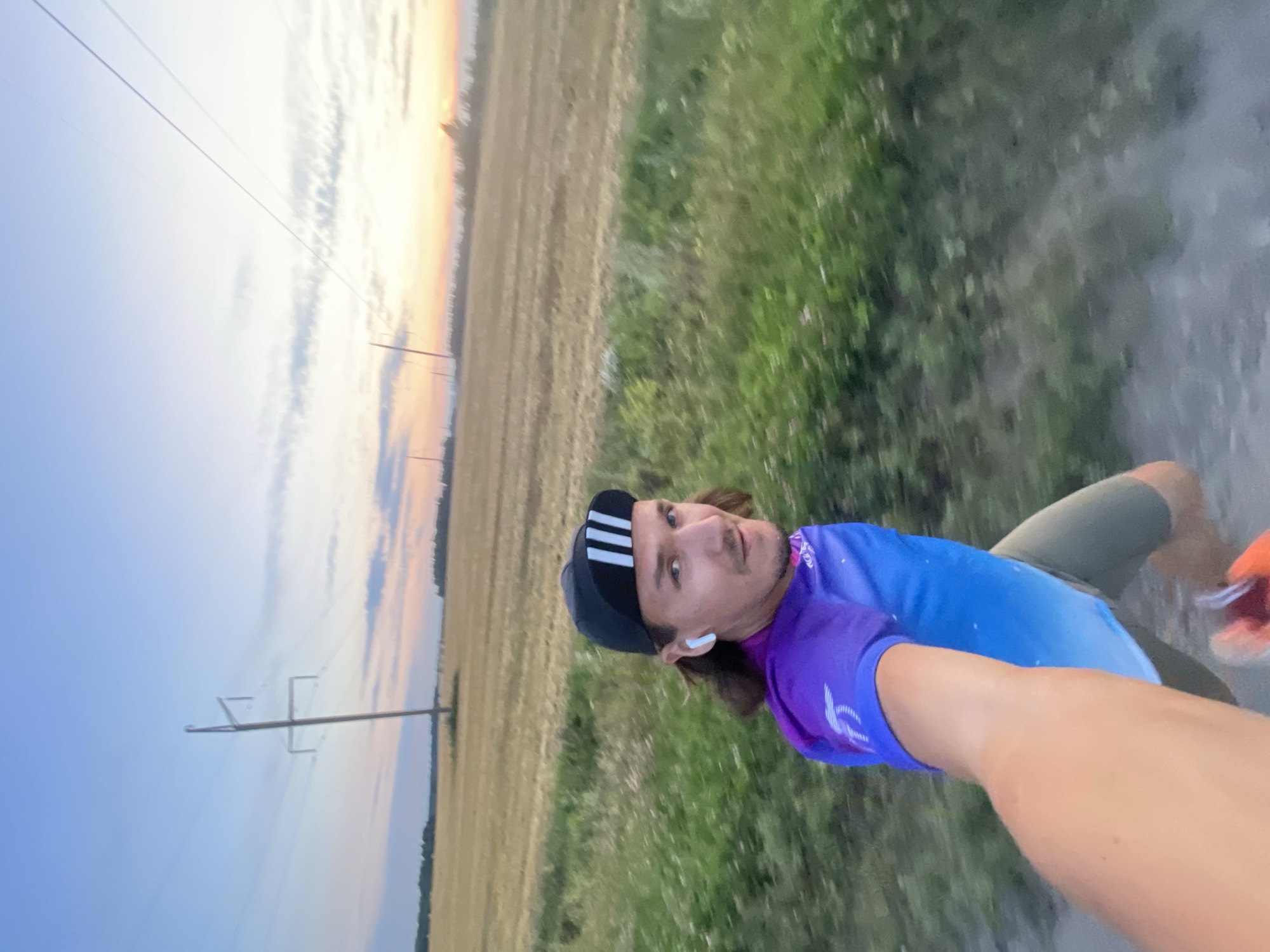 A man with shoulder-length brown hair, wearing a black cap with white stripes and white wireless earbuds, runs towards the camera on a dirt path next to a field of cut hay. He wears a purple and blue gradient t-shirt and olive shorts. The sun is low in the sky, casting a warm glow over the horizon, with power lines visible against a pale blue and pink sky. The scene is set in a rural landscape, possibly on the outskirts of Ol'khovo, Russia, given the presence of agricultural fields and power infrastructure.