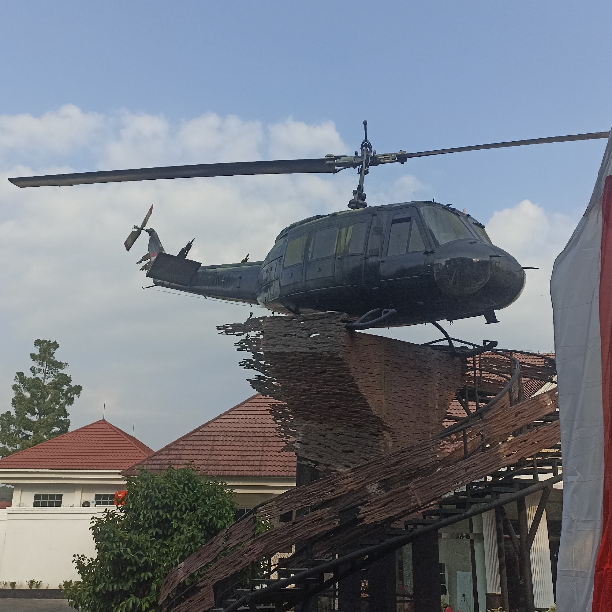 The image shows a dark-colored helicopter mounted on a display structure outdoors. The helicopter appears to be a Vietnam War-era model, possibly a UH-1 Iroquois, commonly known as a "Huey." The helicopter is positioned above a rustic-looking structure made of wood and metal, which features a staircase. In the background, there are buildings with red tile roofs, trees, and a sky with clouds. The scene suggests a monument or a tourist attraction, likely in a public space. The location is identified as Banyurojo, Indonesia. There are no people visible in the frame, and the focus is entirely on the helicopter and its surroundings. A portion of a large Indonesian flag, red and white, is visible on the right side of the frame.
