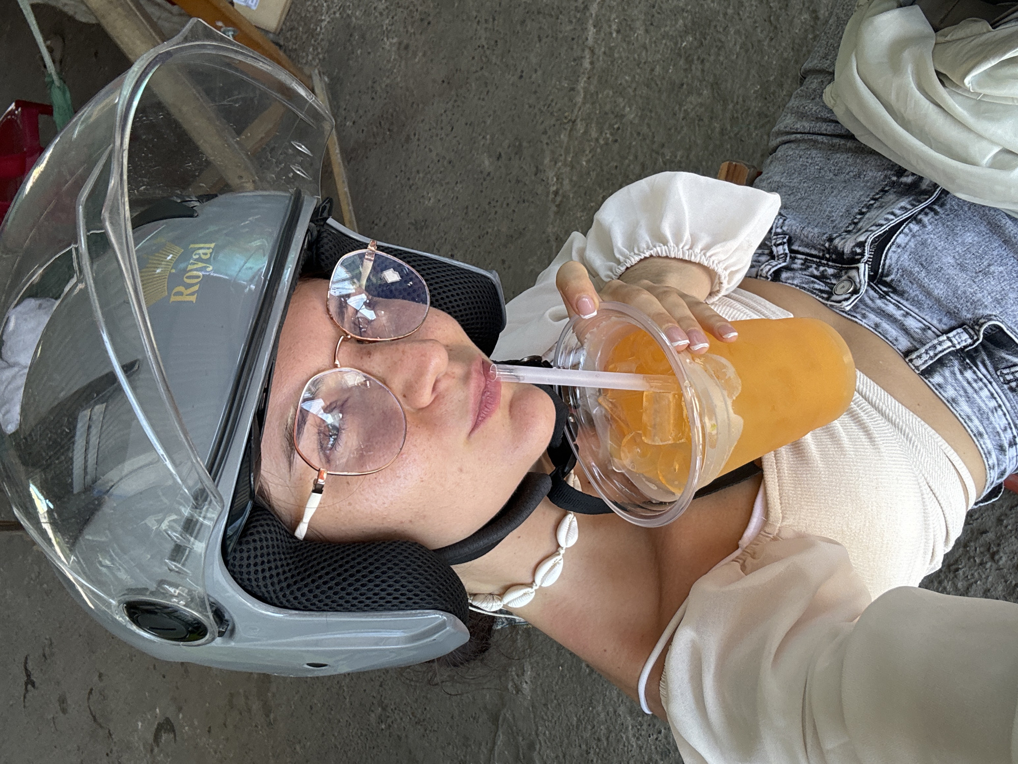 A young woman wearing a grey helmet and glasses is taking a selfie while holding a plastic cup of orange juice with ice and a straw. She is also wearing a white top and denim shorts. The background appears to be an outdoor setting with a concrete ground and some blurred objects in the distance, possibly a street scene in Hồng Sơn, Vietnam.