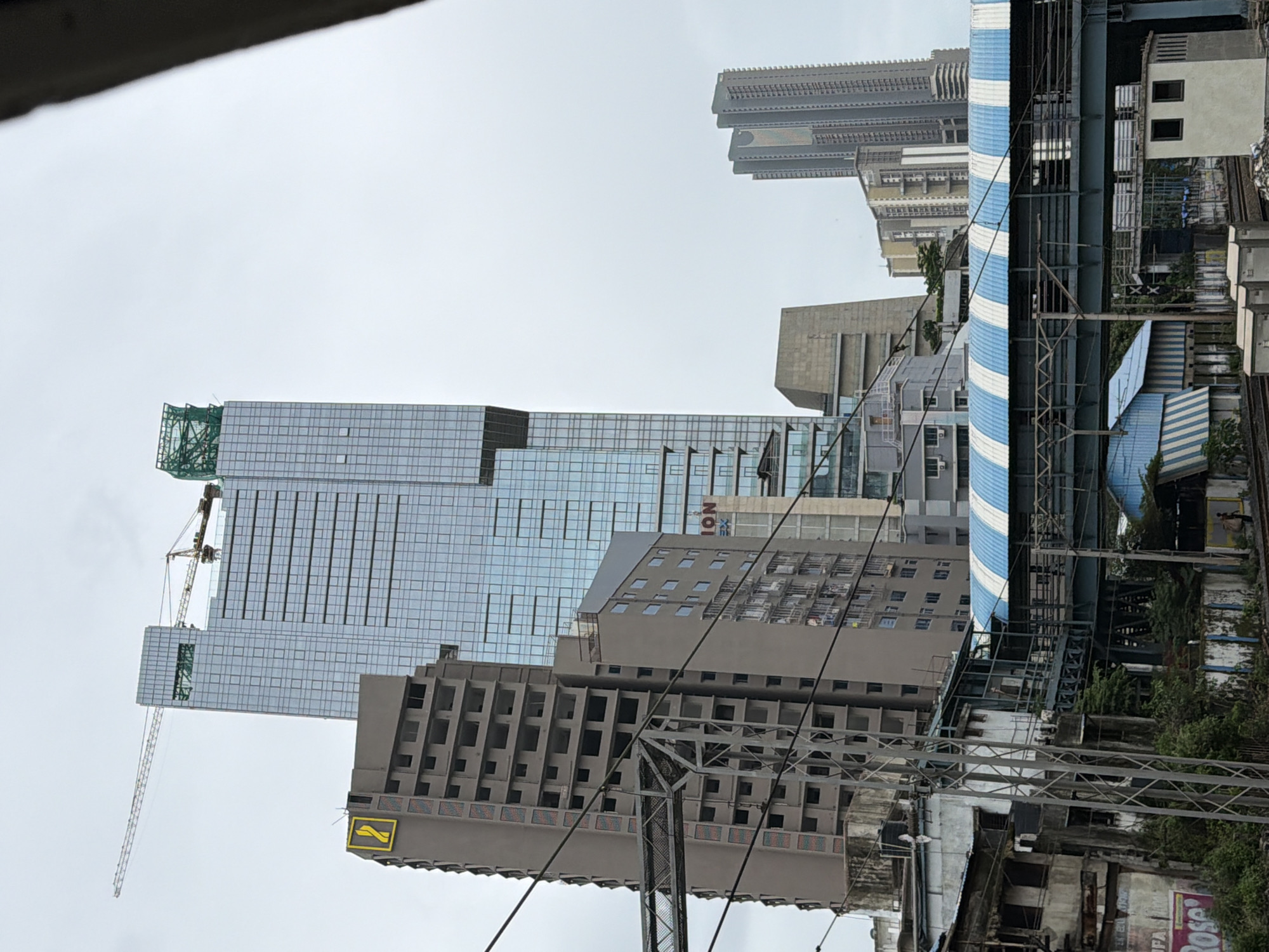 This image shows a view of the cityscape of Mumbai, India, with a focus on modern skyscrapers and older, unfinished buildings. In the foreground, there is a pedestrian bridge with a blue and white striped roof, crossing over railway tracks. Various utility poles and wires are visible.
Behind the bridge, several buildings rise up. To the left, a tall, gray building appears to be under construction, with exposed concrete floors and windows missing. It features a yellow logo with a stylized '2'. Adjacent to this, and slightly behind, is a very tall, sleek, glass-covered skyscraper, which is clearly a modern office or residential building. A construction crane is visible on top of this skyscraper, indicating ongoing development.
Further in the background, other modern high-rise buildings of varying designs and heights are scattered across the skyline. The sky is overcast, suggesting a cloudy day. The scene depicts a blend of old and new architecture, characteristic of rapidly developing urban environments like Mumbai, with a clear contrast between ongoing construction, modern commercial structures, and existing infrastructure.