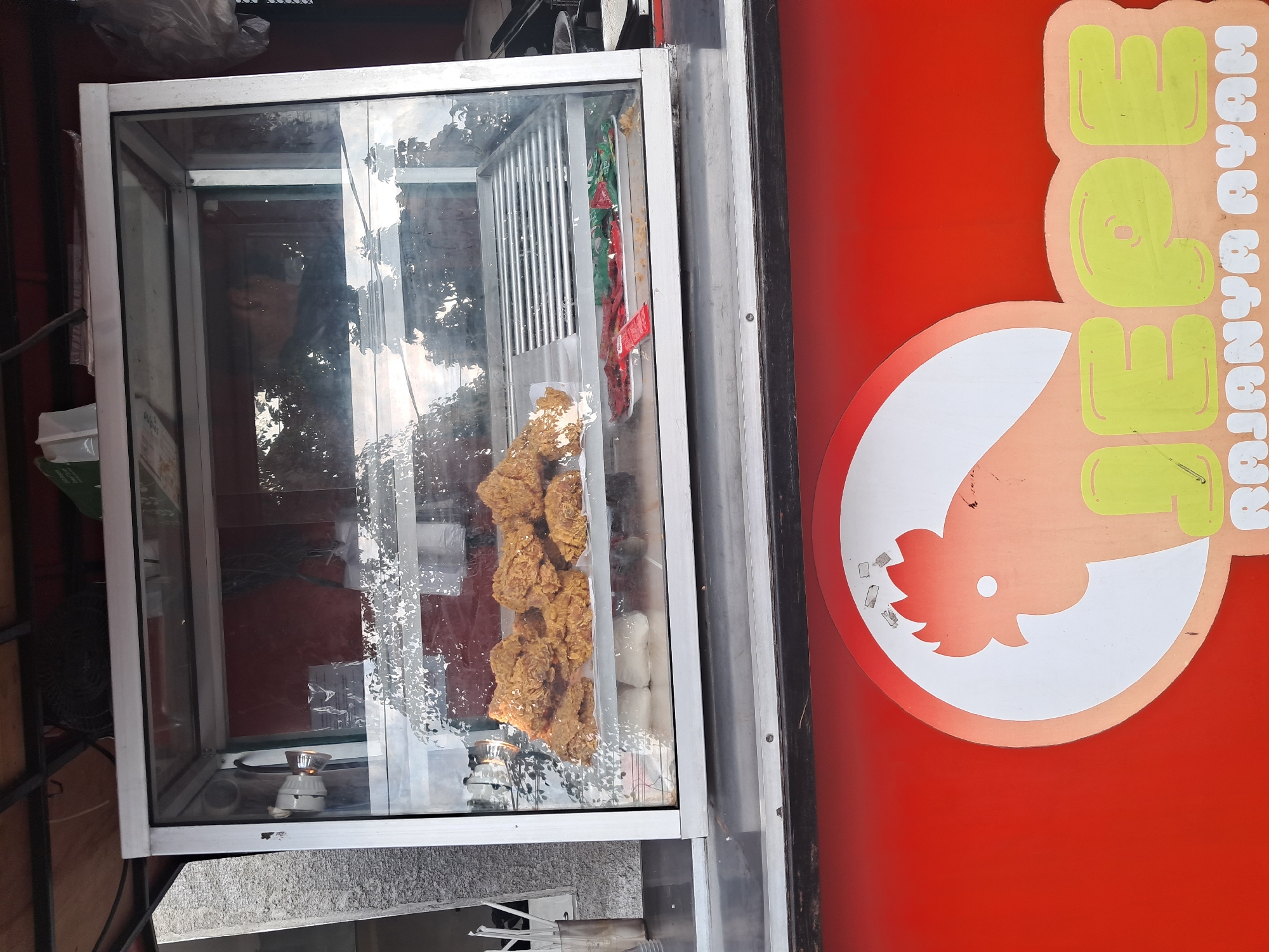 A storefront display case on a red food cart in Klaten, Indonesia, contains fried chicken. The display case has a silver frame and glass front and top. Inside, on the top shelf, are several pieces of golden-brown fried chicken. On the bottom shelf, there are packets of green and red condiments, and some white rectangular items underneath the chicken. The food cart itself is red and features a circular logo of a stylized chicken head and the text "JEPE RAJAnya AYAM" in yellow and white lettering.