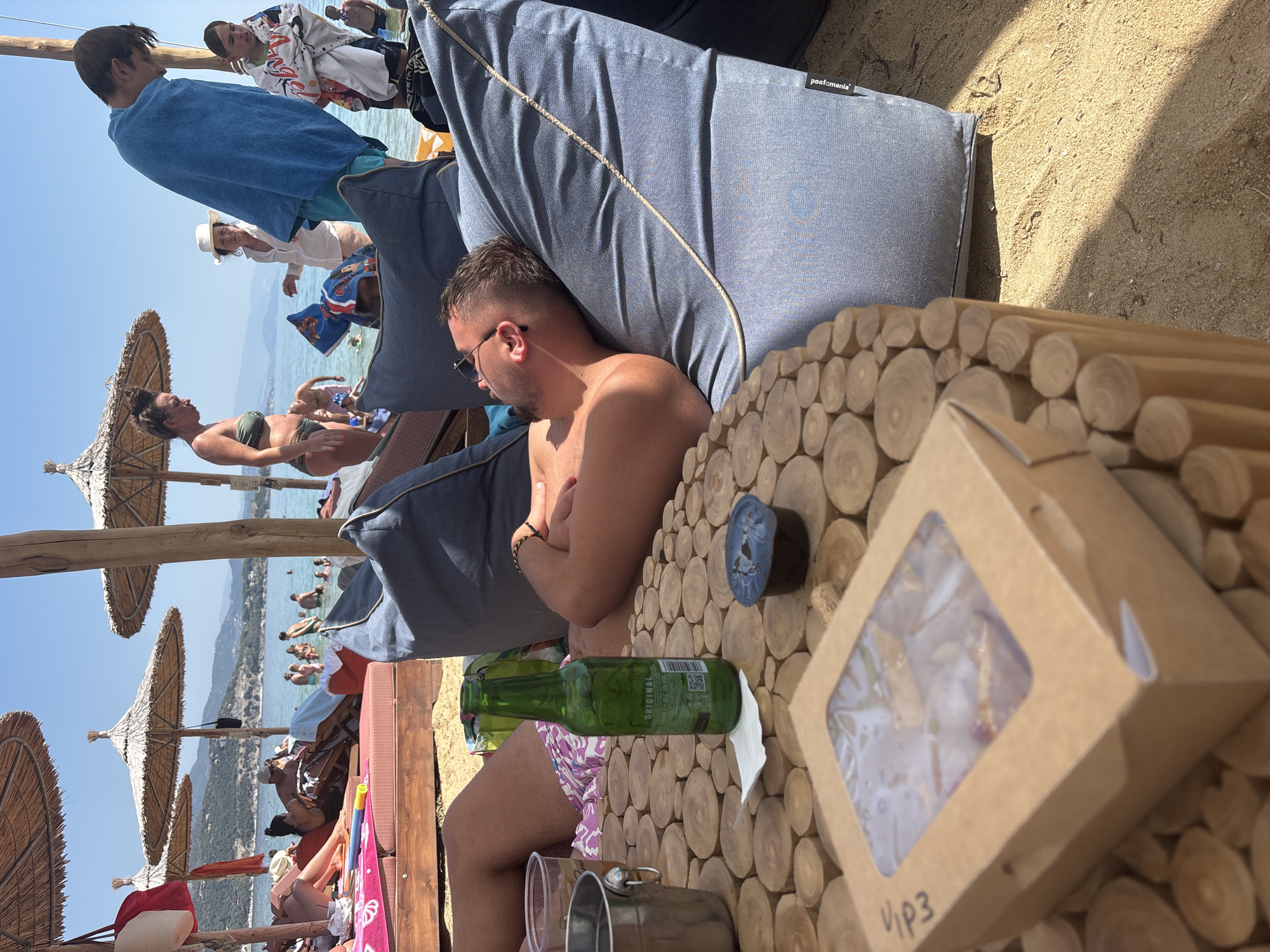 A man is lying on a large, blue beanbag chair on a sandy beach in Nea Irakleitsa, Greece. He is wearing sunglasses and has his arms crossed. In front of him, on a circular wooden table made of cut logs, is a green bottle of beer, a small plastic container with a dark substance, and a cardboard box with a clear window. Behind him, other people are visible on the beach, some in the water and others under straw umbrellas. The water is calm and blue, and the sky is clear.