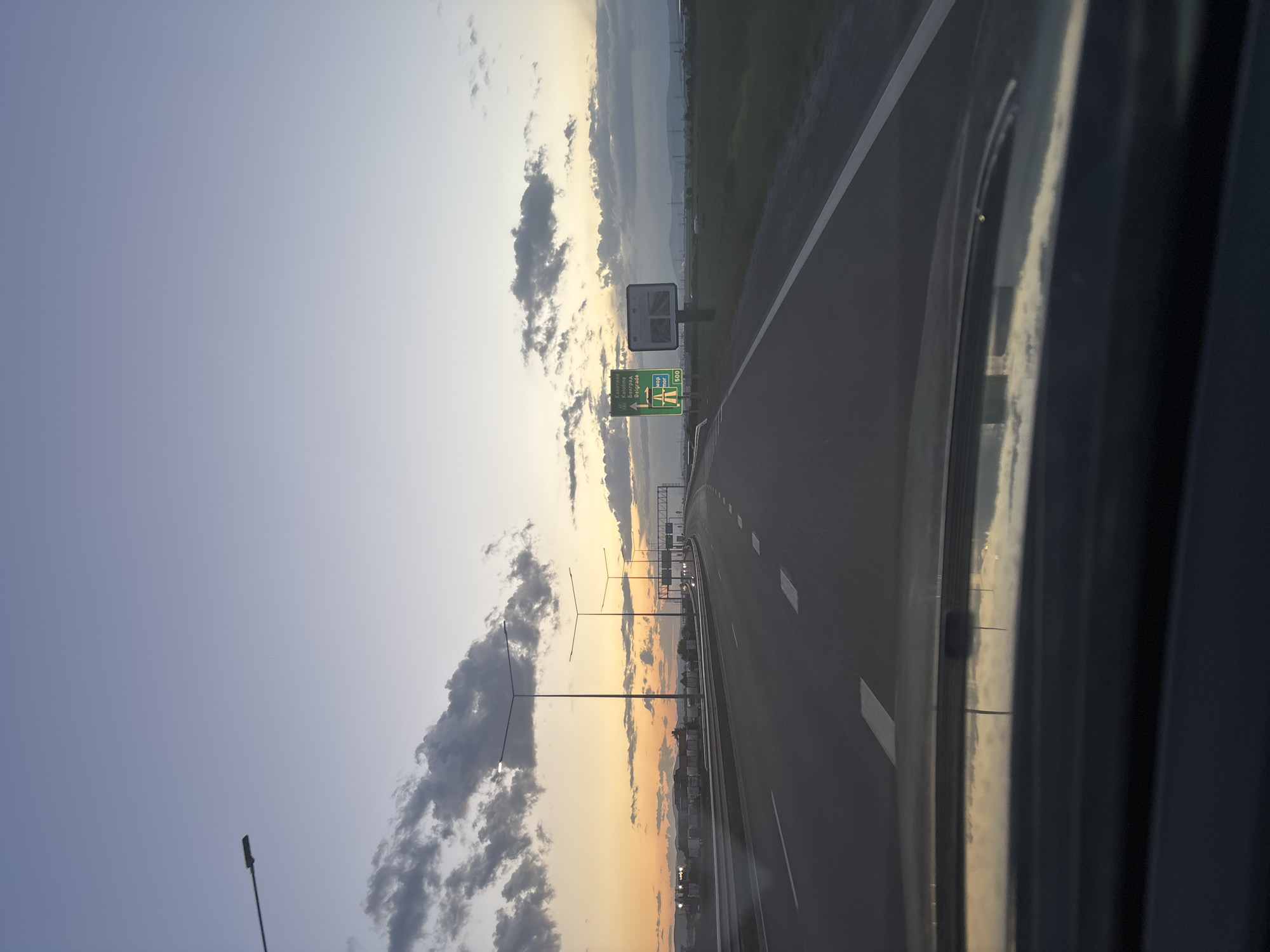 The image shows a highway leading into Sofia, Bulgaria, at sunset. The road has multiple lanes and is marked with white lines. Streetlights line the road, and a large sign indicates directions to Belgrade. The sky is a gradient of blues, oranges, and grays with scattered clouds. The view appears to be taken from inside a vehicle.